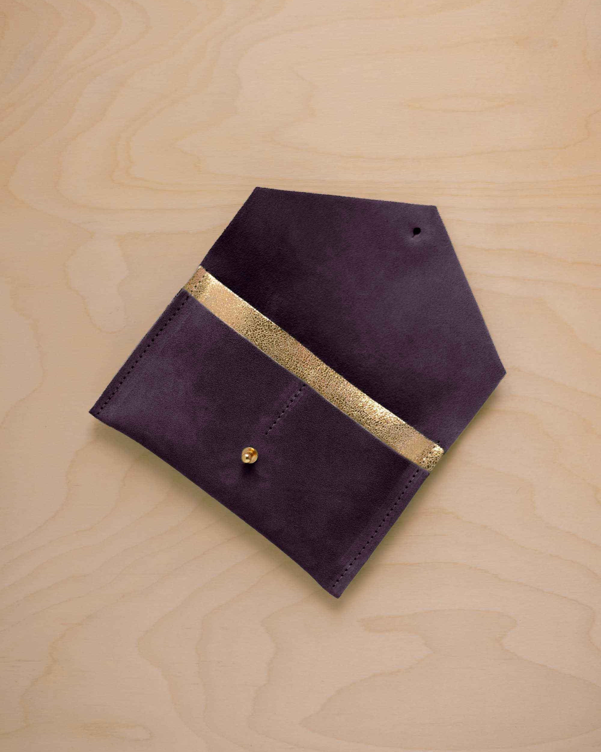 A blackcurrant dark purple suede pouch with a metallic gold contrast pocket and a small gold stud, displayed on a light wood grain background.