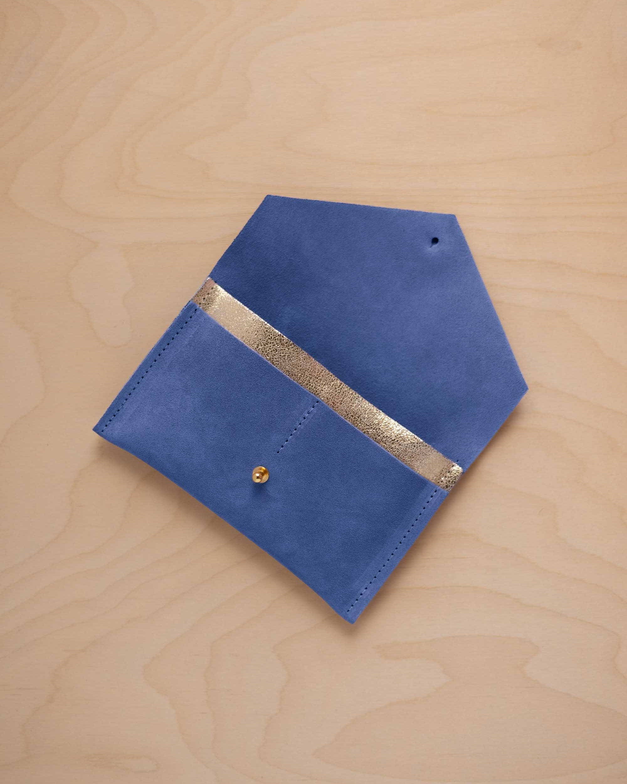 A cobalt blue suede pouch with a metallic gold contrast pocket and a small gold stud, displayed on a light wood grain background.