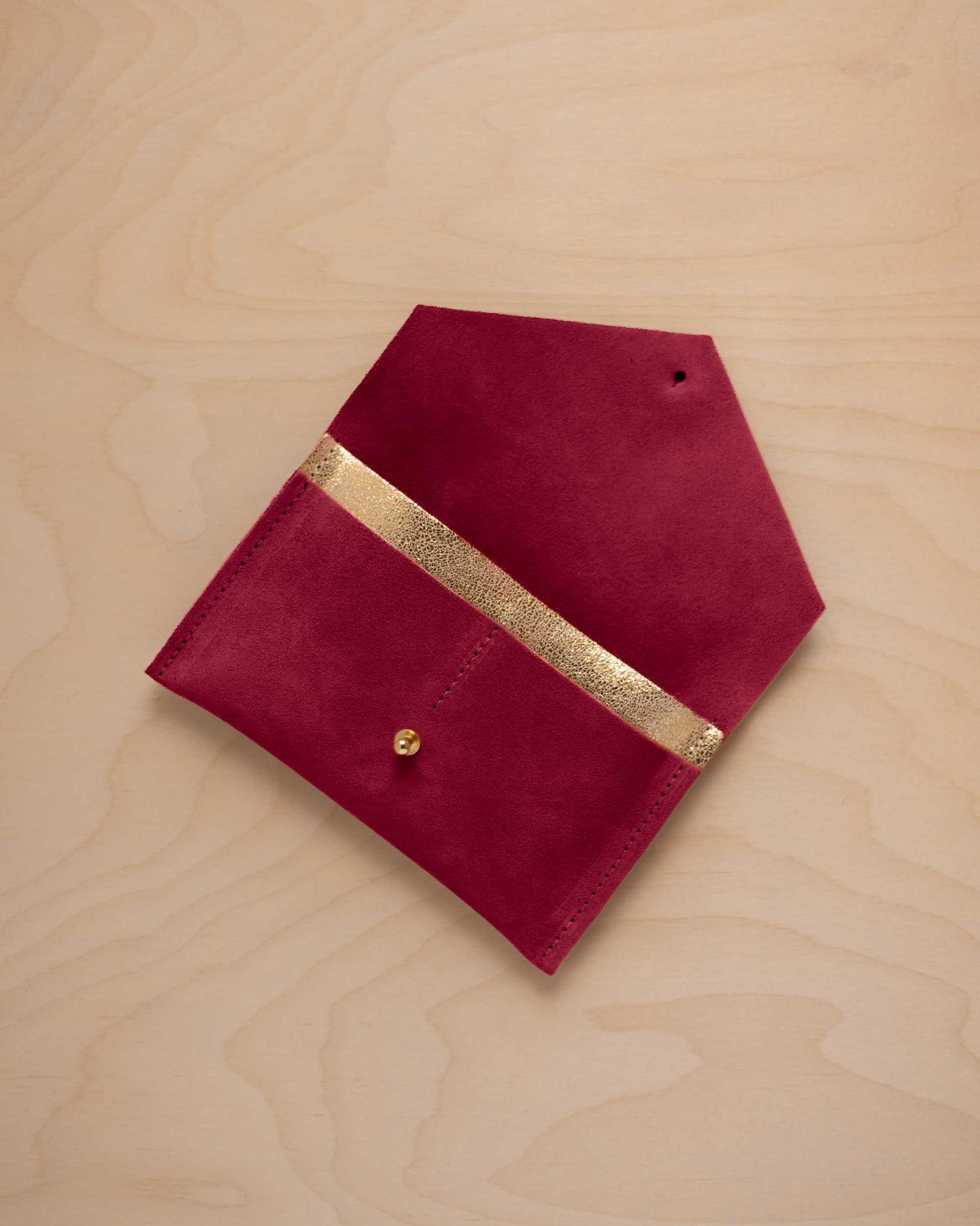 A crimson red suede pouch with a metallic gold contrast pocket and a small gold stud, displayed on a light wood grain background.