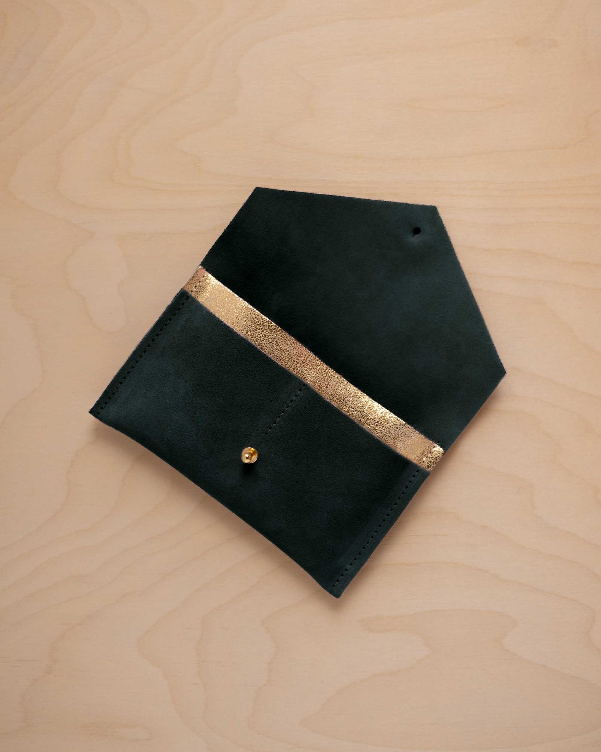 A deep forest green coloured suede pouch with a metallic gold contrast pocket and a small gold stud, displayed on a light wood grain background.
