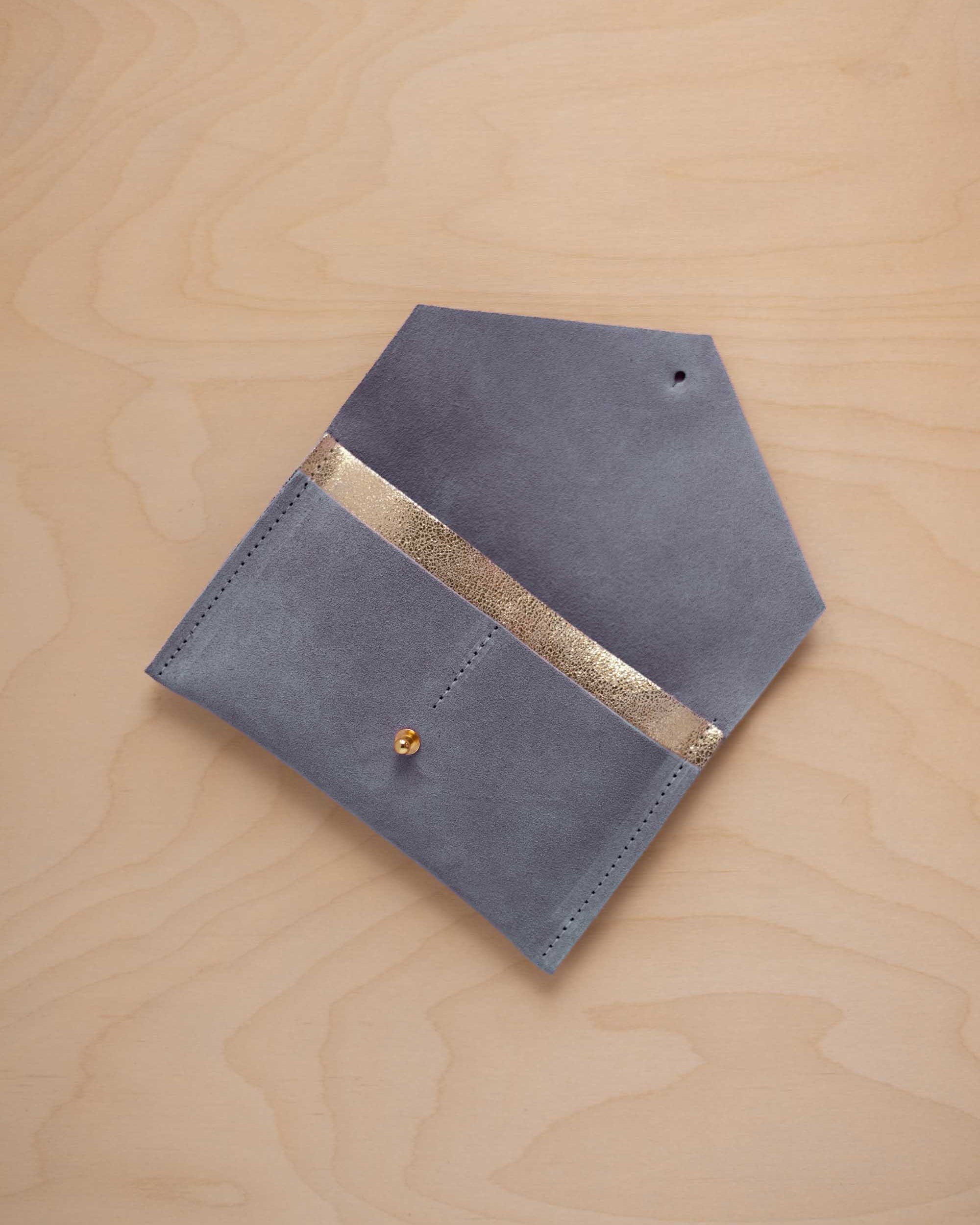 A wdusky blue suede pouch with a metallic gold contrast pocket and a small gold stud, displayed on a light wood grain background.