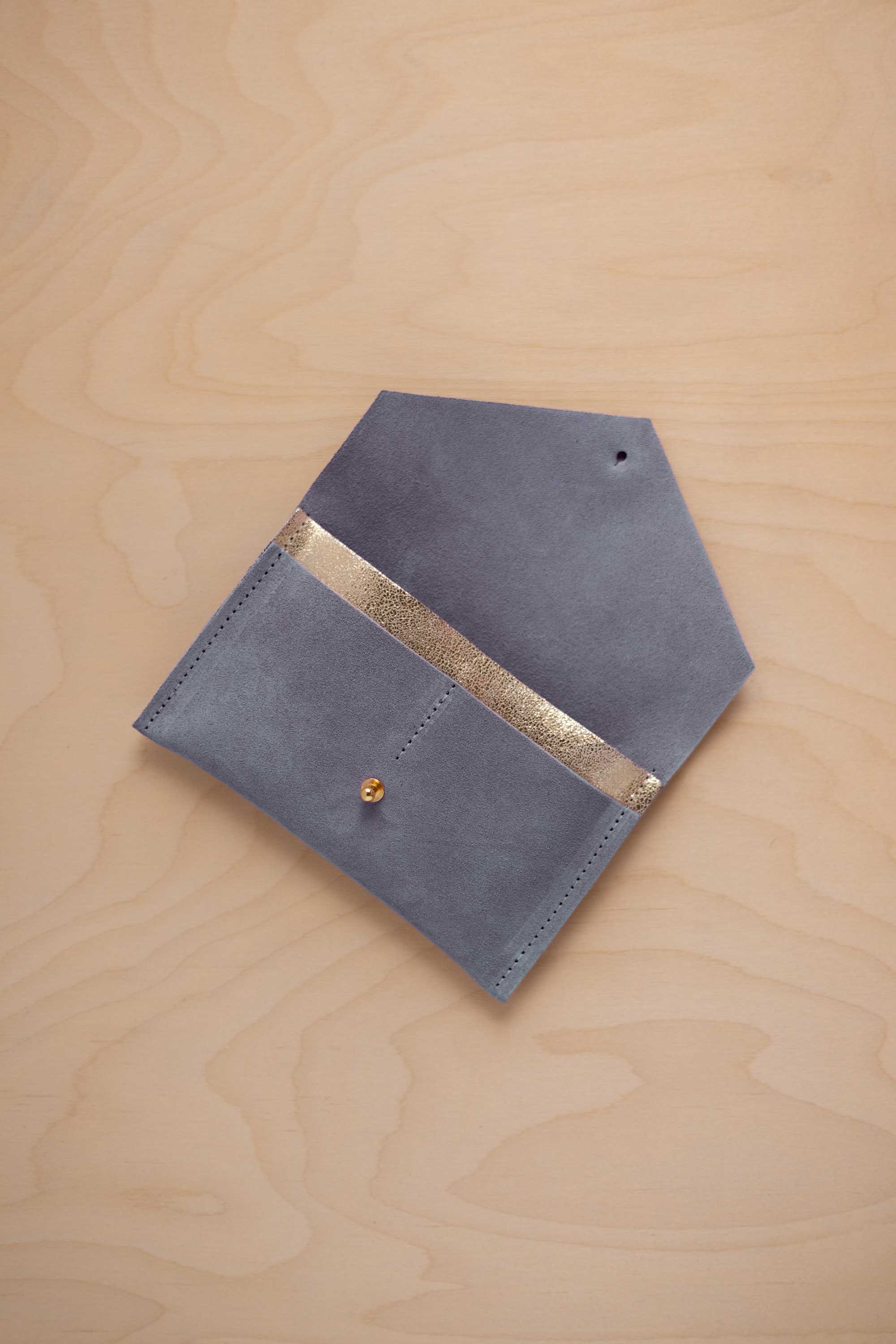 A wdusky blue suede pouch with a metallic gold contrast pocket and a small gold stud, displayed on a light wood grain background.