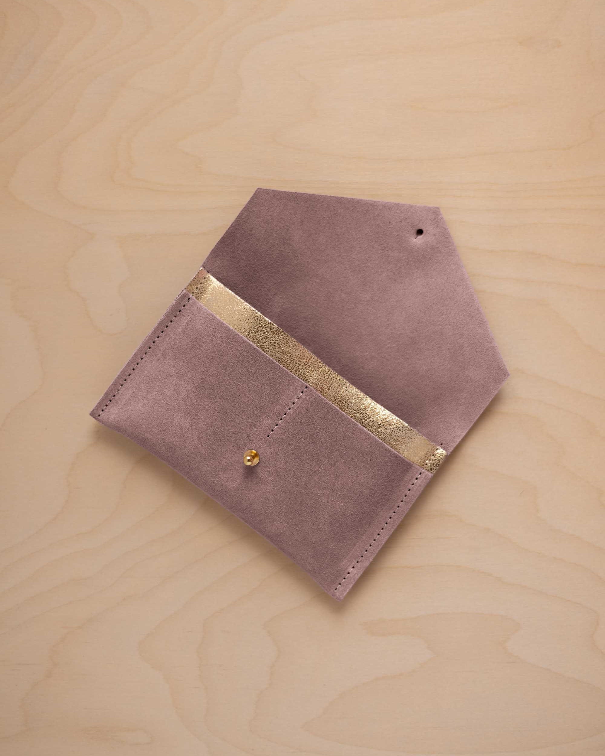 A dusty mauve grey suede pouch with a metallic gold contrast pocket and a small gold stud, displayed on a light wood grain background.