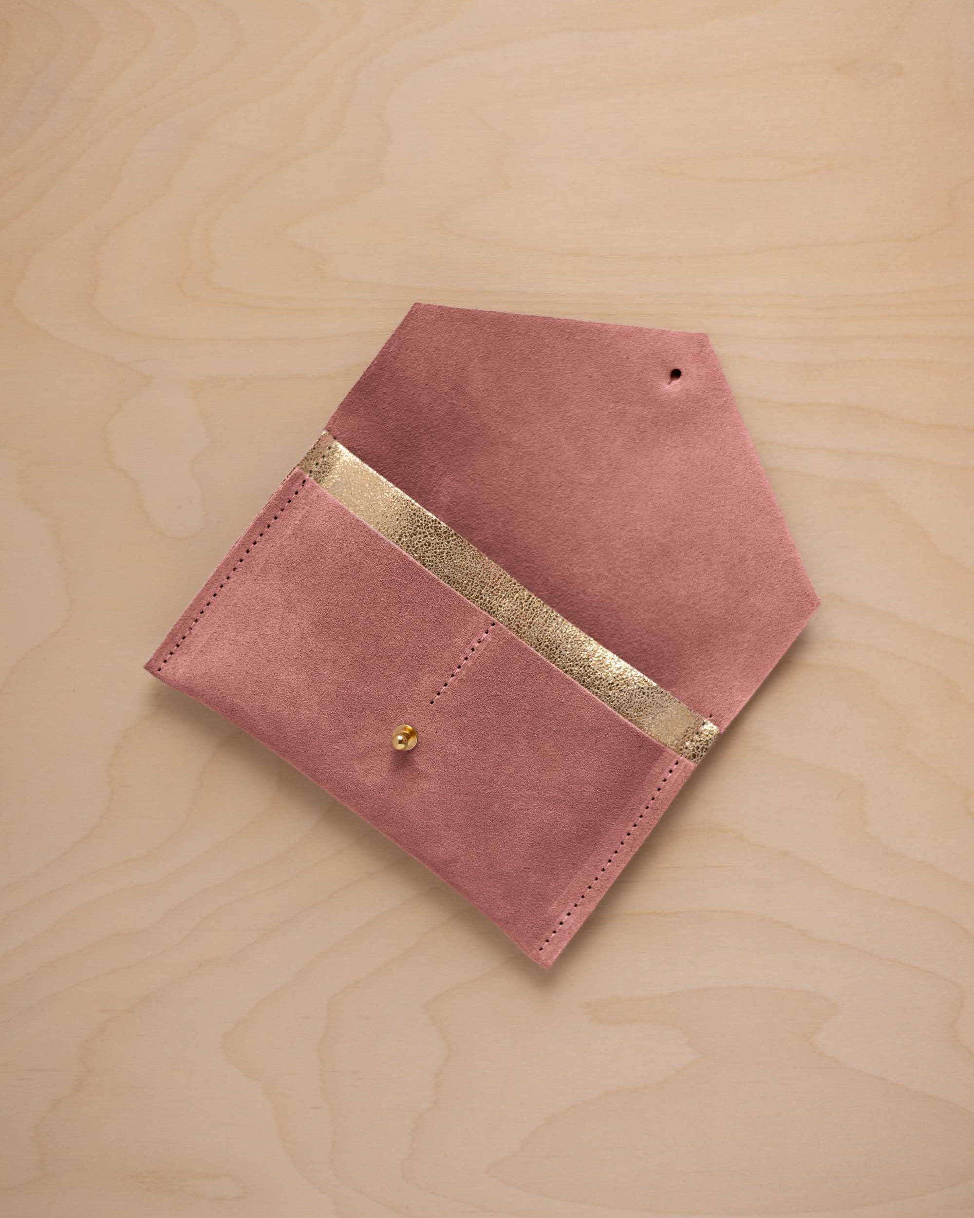 A dusty rose pink coloured suede pouch with a metallic gold contrast pocket and a small gold stud, displayed on a light wood grain background.
