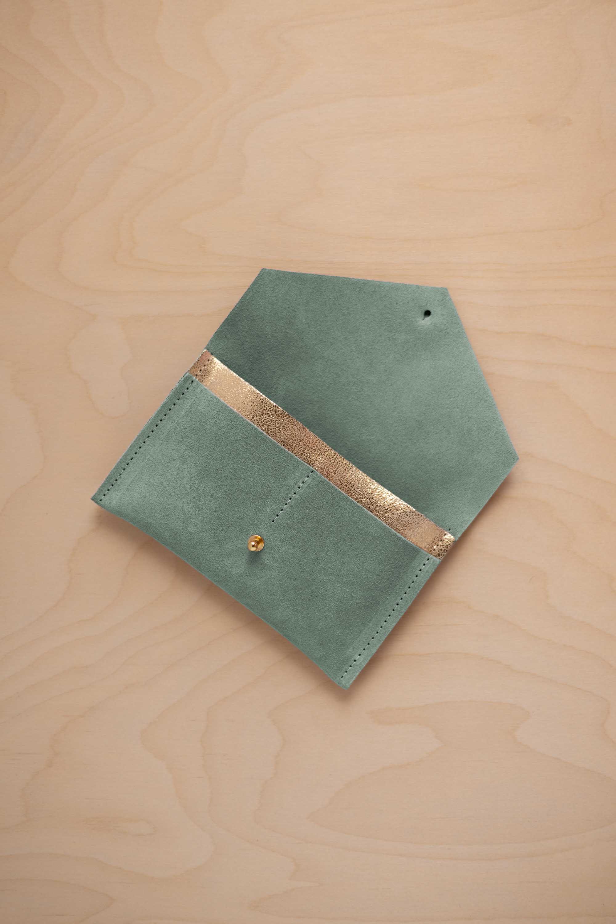A eucalyptus sage green coloured suede pouch with a metallic gold contrast pocket and a small gold stud, displayed on a light wood grain background.