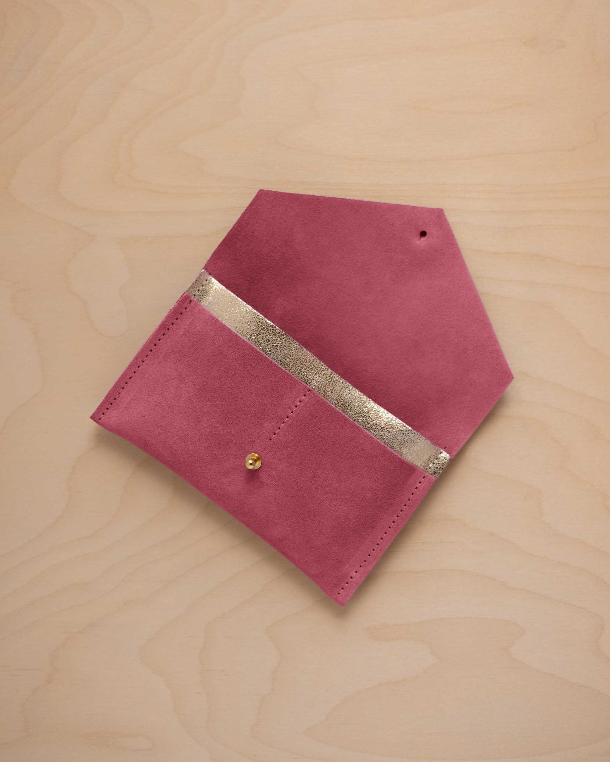 A faded rose pink suede pouch with a metallic gold contrast pocket and a small gold stud, displayed on a light wood grain background.