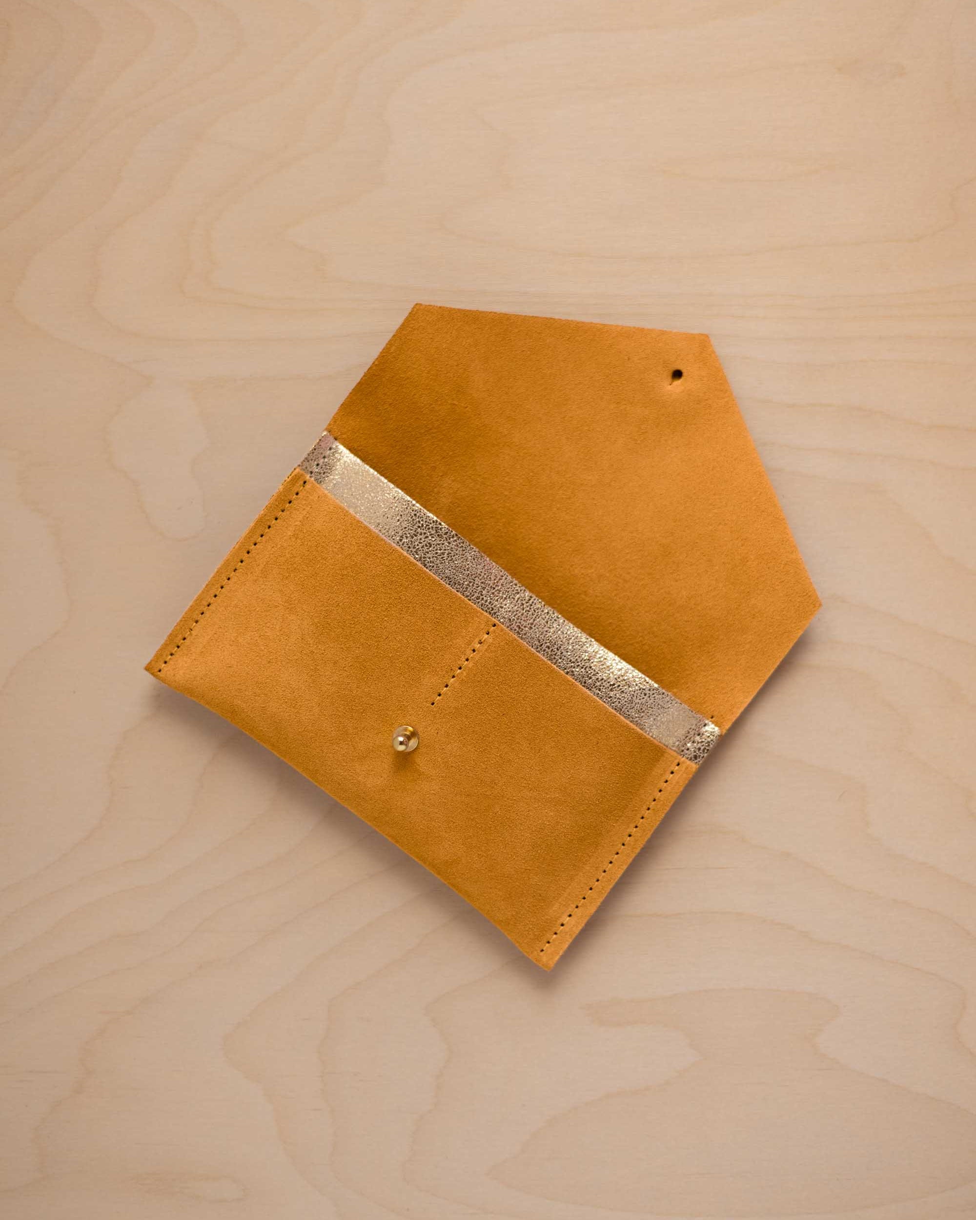 A golden ochre mustard yellow suede pouch with a metallic gold contrast pocket and a small gold stud, displayed on a light wood grain background.