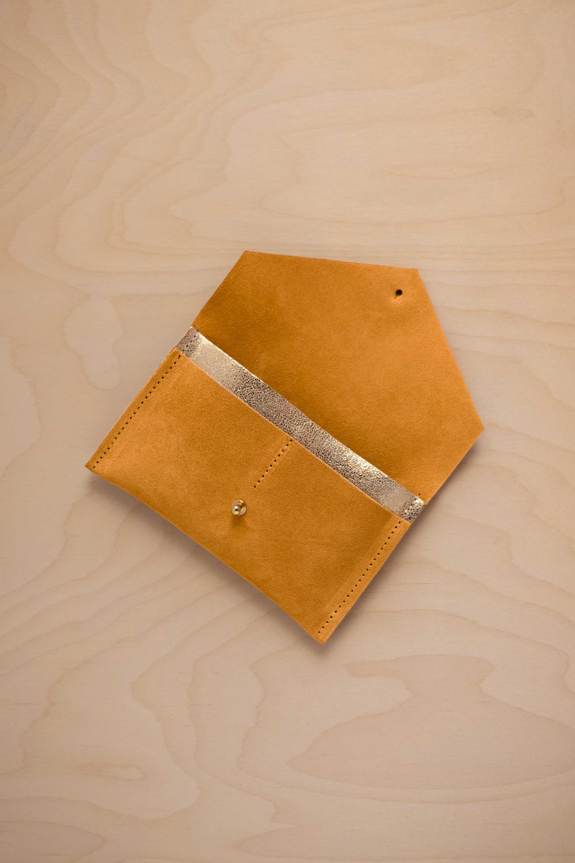 A golden ochre mustard yellow suede pouch with a metallic gold contrast pocket and a small gold stud, displayed on a light wood grain background.
