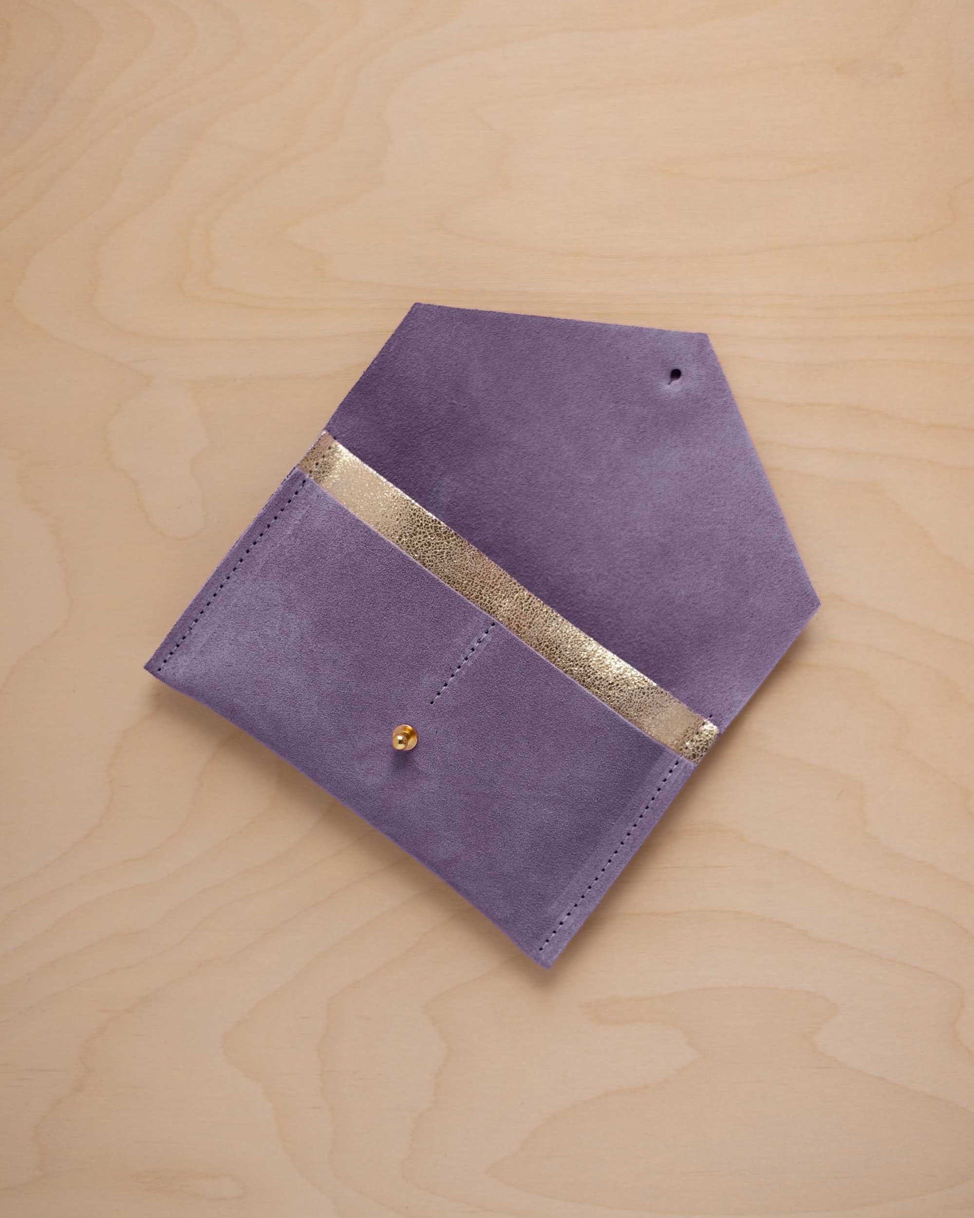A lavender suede pouch with a metallic gold contrast pocket and a small gold stud, displayed on a light wood grain background.