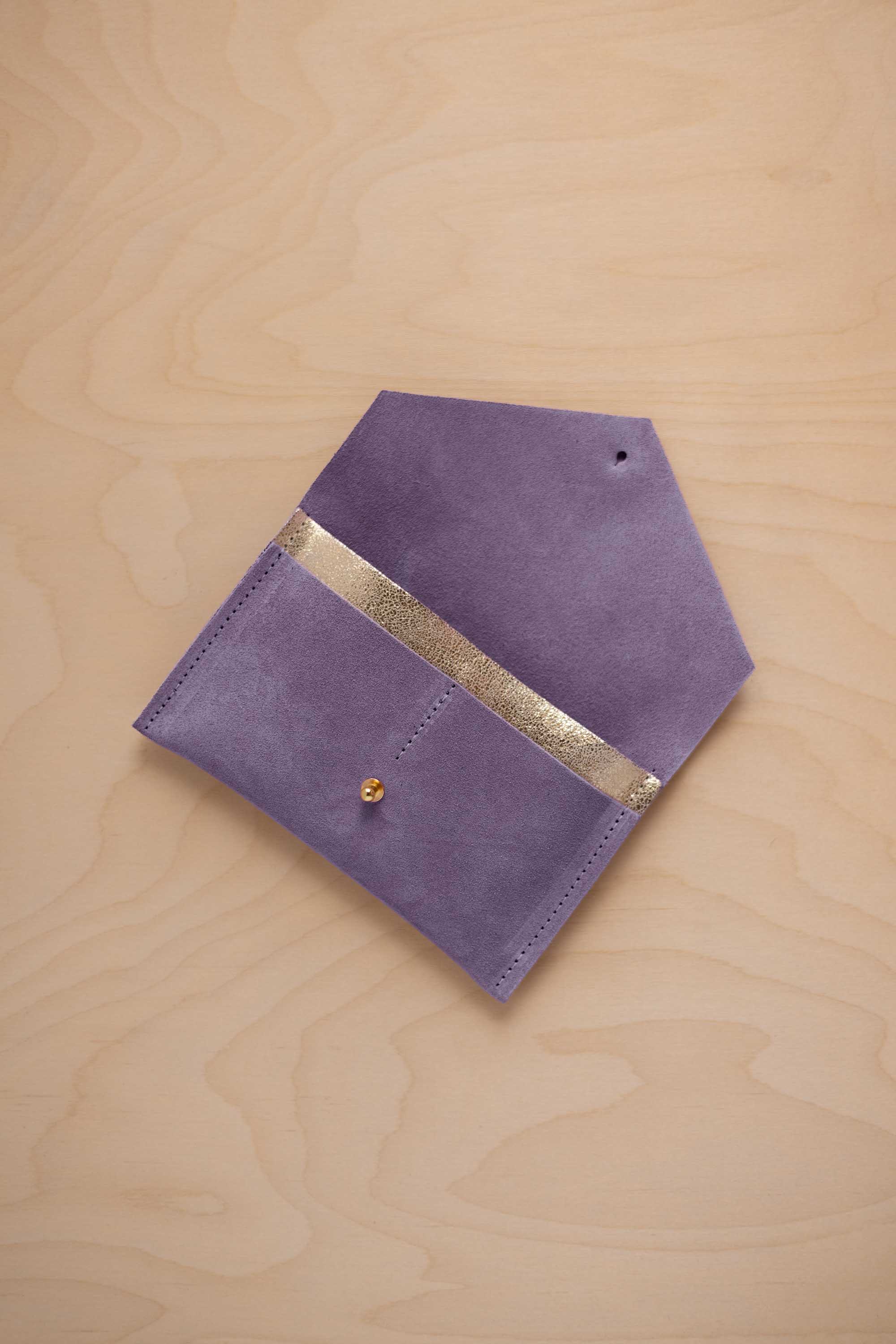 A lavender suede pouch with a metallic gold contrast pocket and a small gold stud, displayed on a light wood grain background.