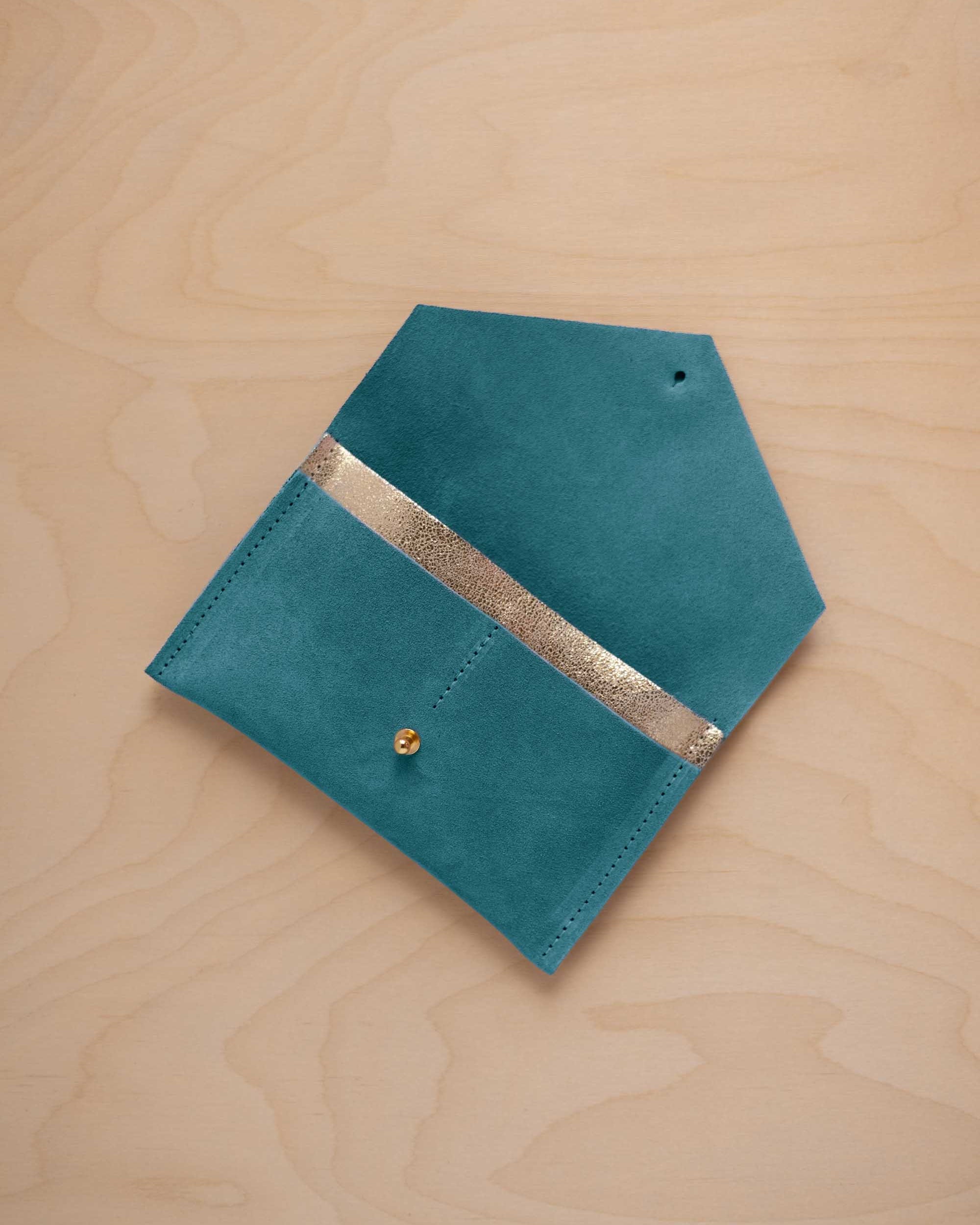 A marine teal suede pouch with a metallic gold contrast pocket and a small gold stud, displayed on a light wood grain background.