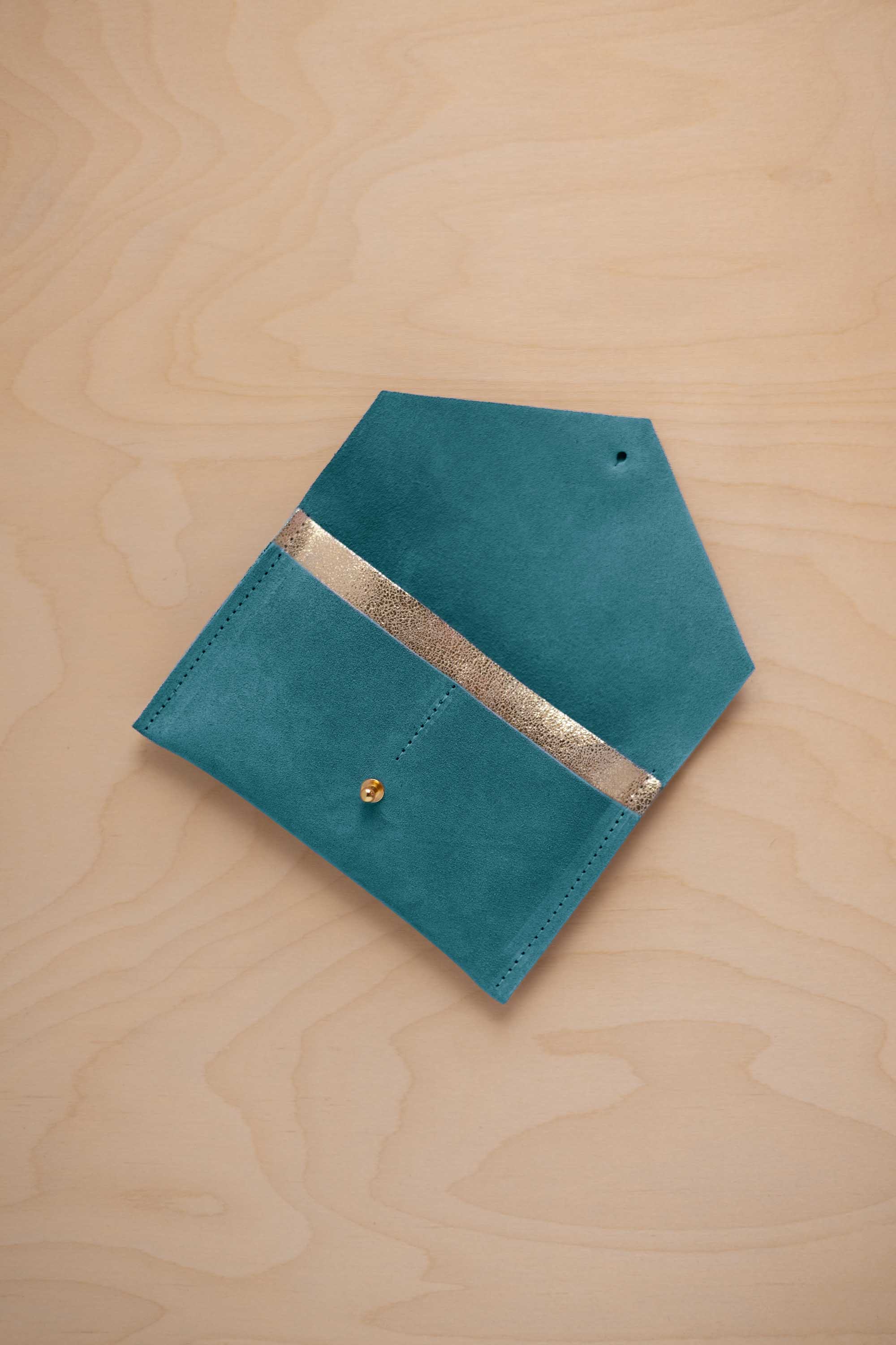 A marine teal suede pouch with a metallic gold contrast pocket and a small gold stud, displayed on a light wood grain background.