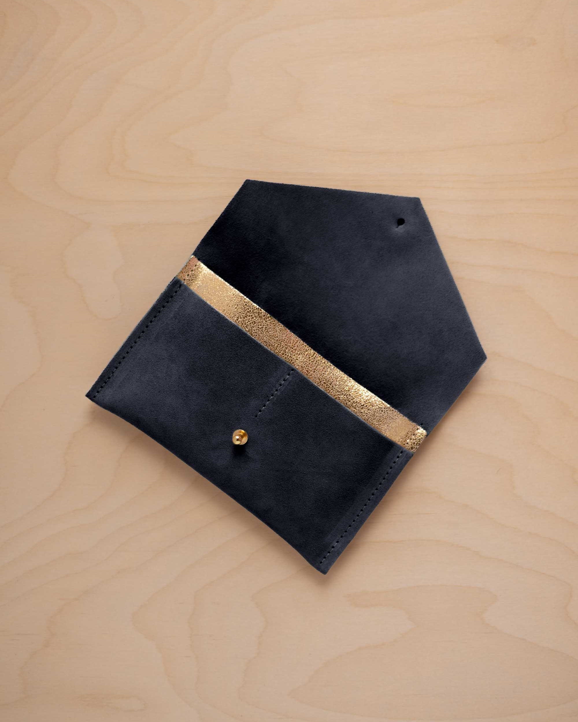 A midnight navy blue suede pouch with a metallic gold contrast pocket and a small gold stud, displayed on a light wood grain background.