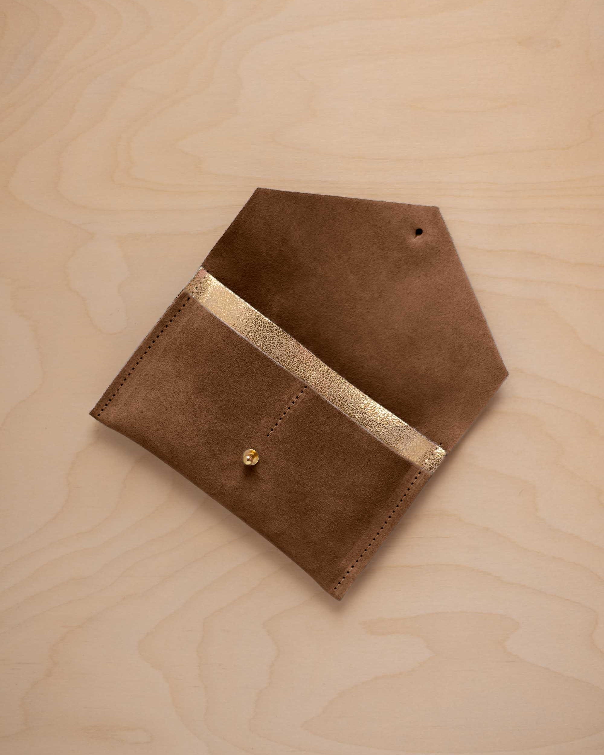 A mocha brown suede pouch with a metallic gold contrast pocket and a small gold stud, displayed on a light wood grain background.