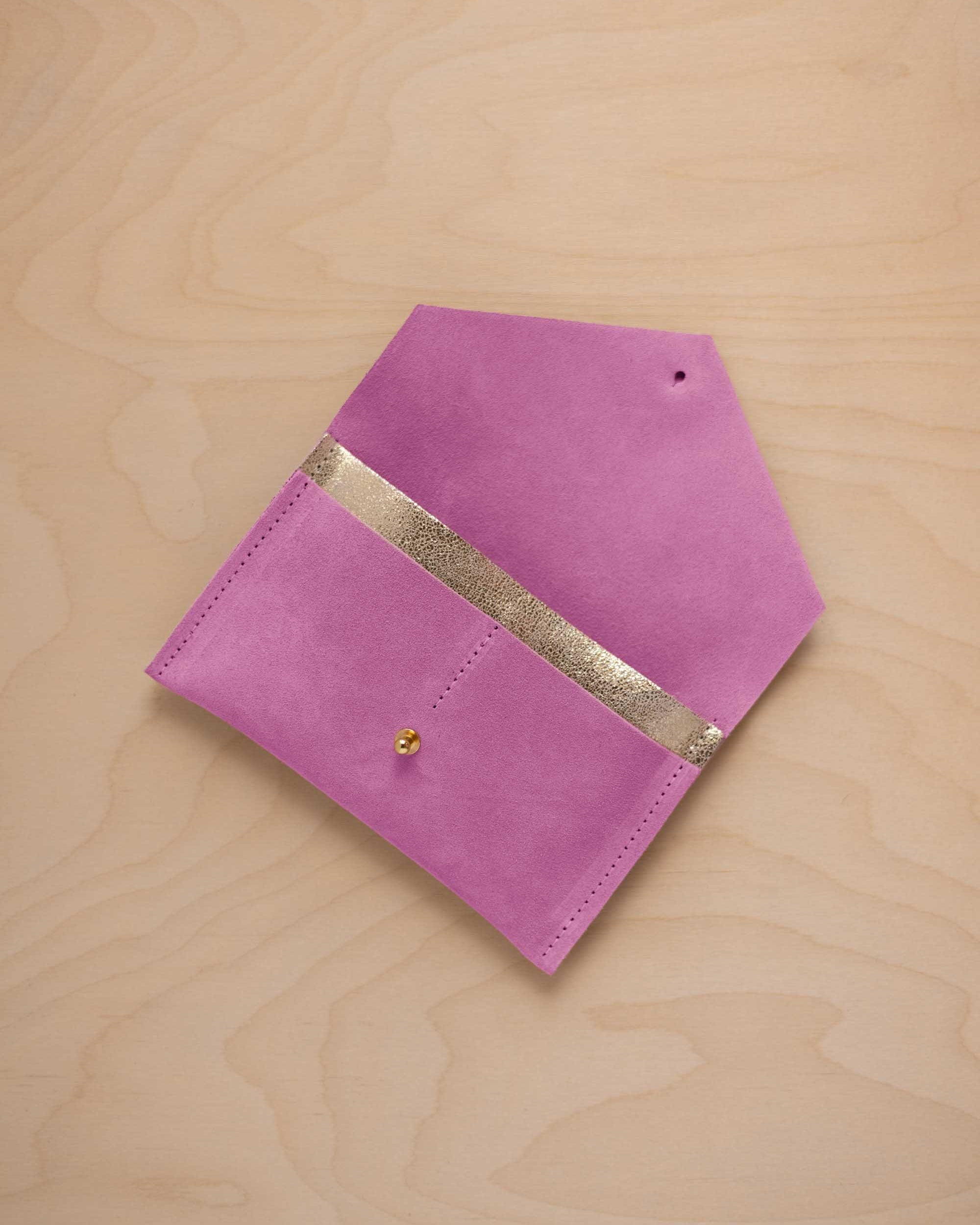 A peony pink suede pouch with a metallic gold contrast pocket and a small gold stud, displayed on a light wood grain background.