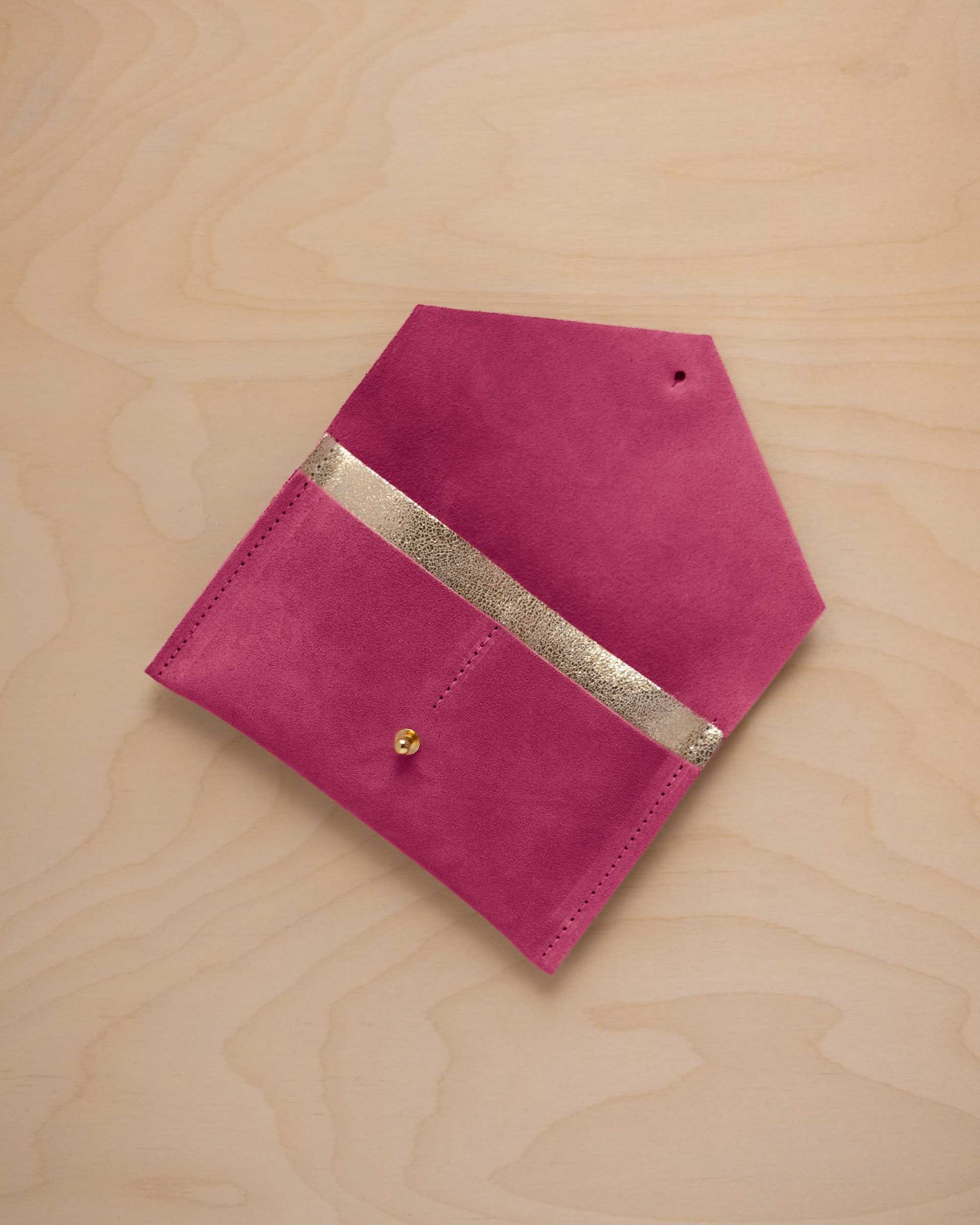 A raspberry pink suede pouch with a metallic gold contrast pocket and a small gold stud, displayed on a light wood grain background.