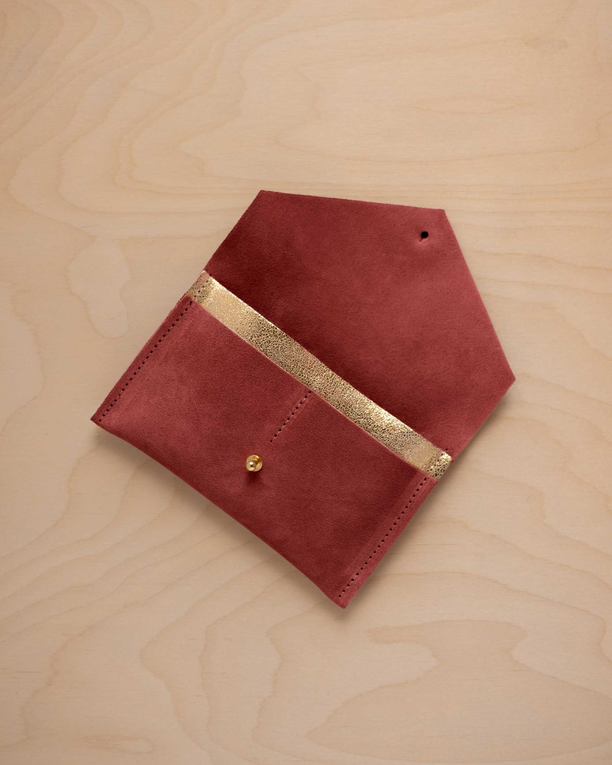 A rust red suede pouch with a metallic gold contrast pocket and a small gold stud, displayed on a light wood grain background.