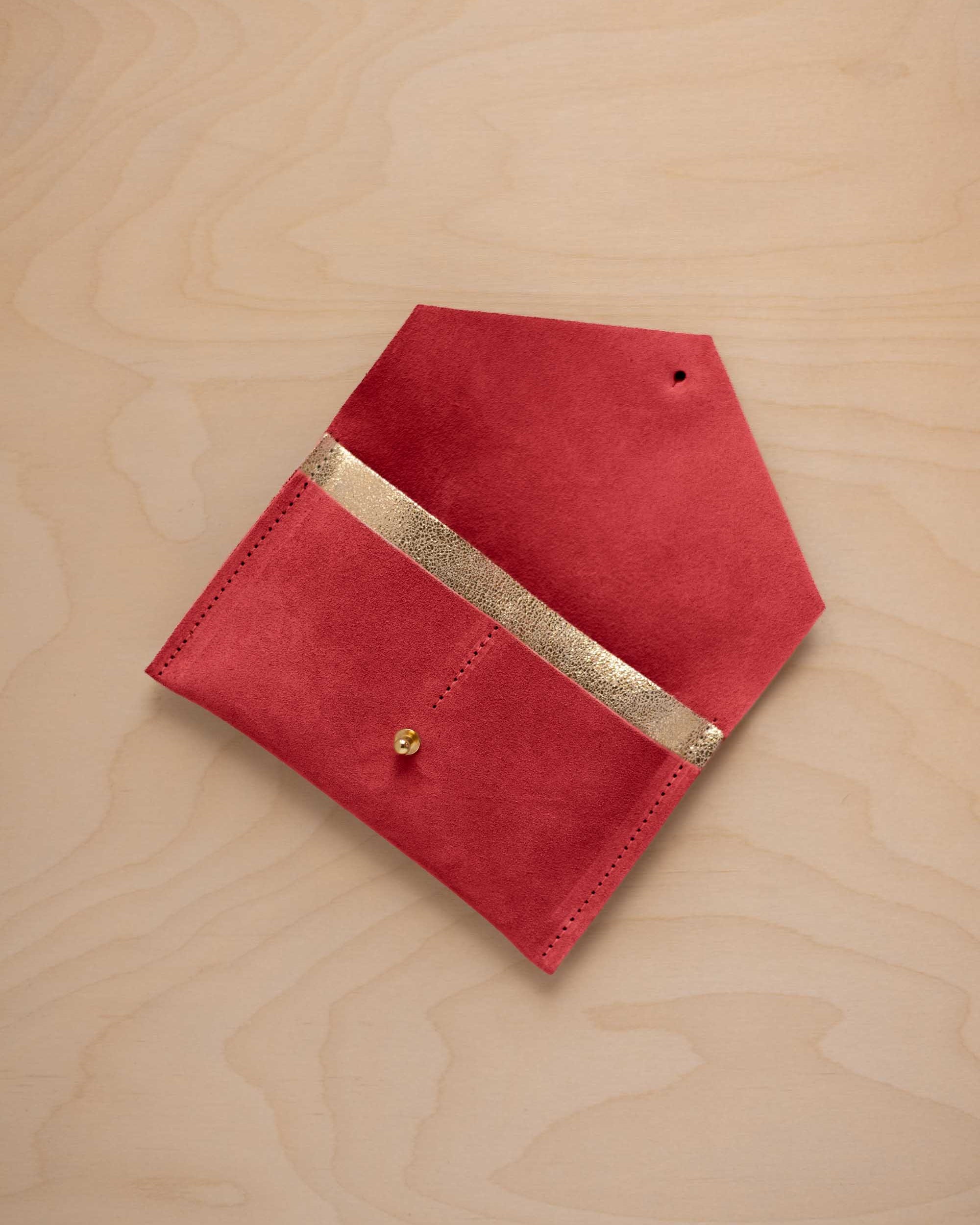 A scarlet red suede pouch with a metallic gold contrast pocket and a small gold stud, displayed on a light wood grain background.