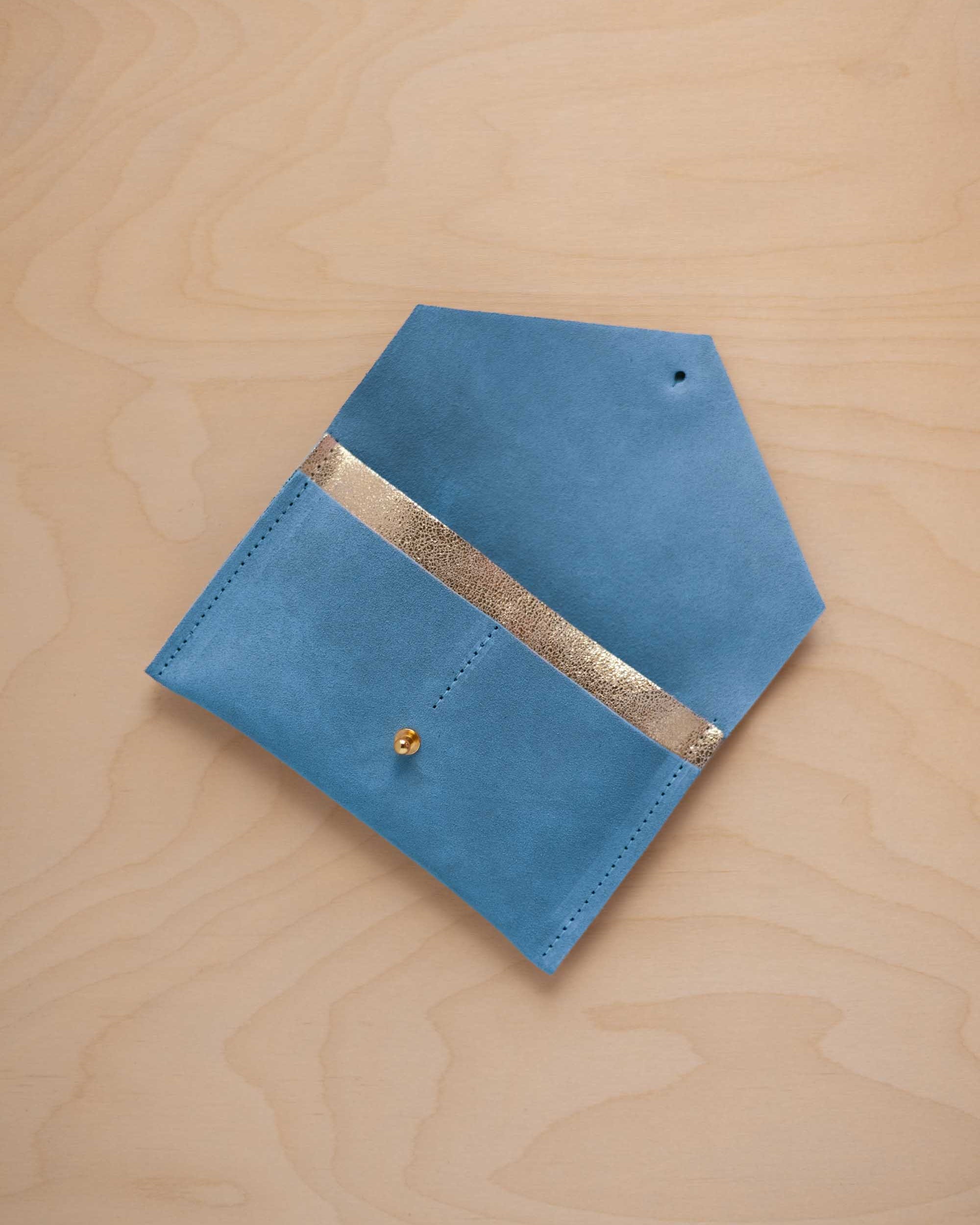 A sky blue suede pouch with a metallic gold contrast pocket and a small gold stud, displayed on a light wood grain background.