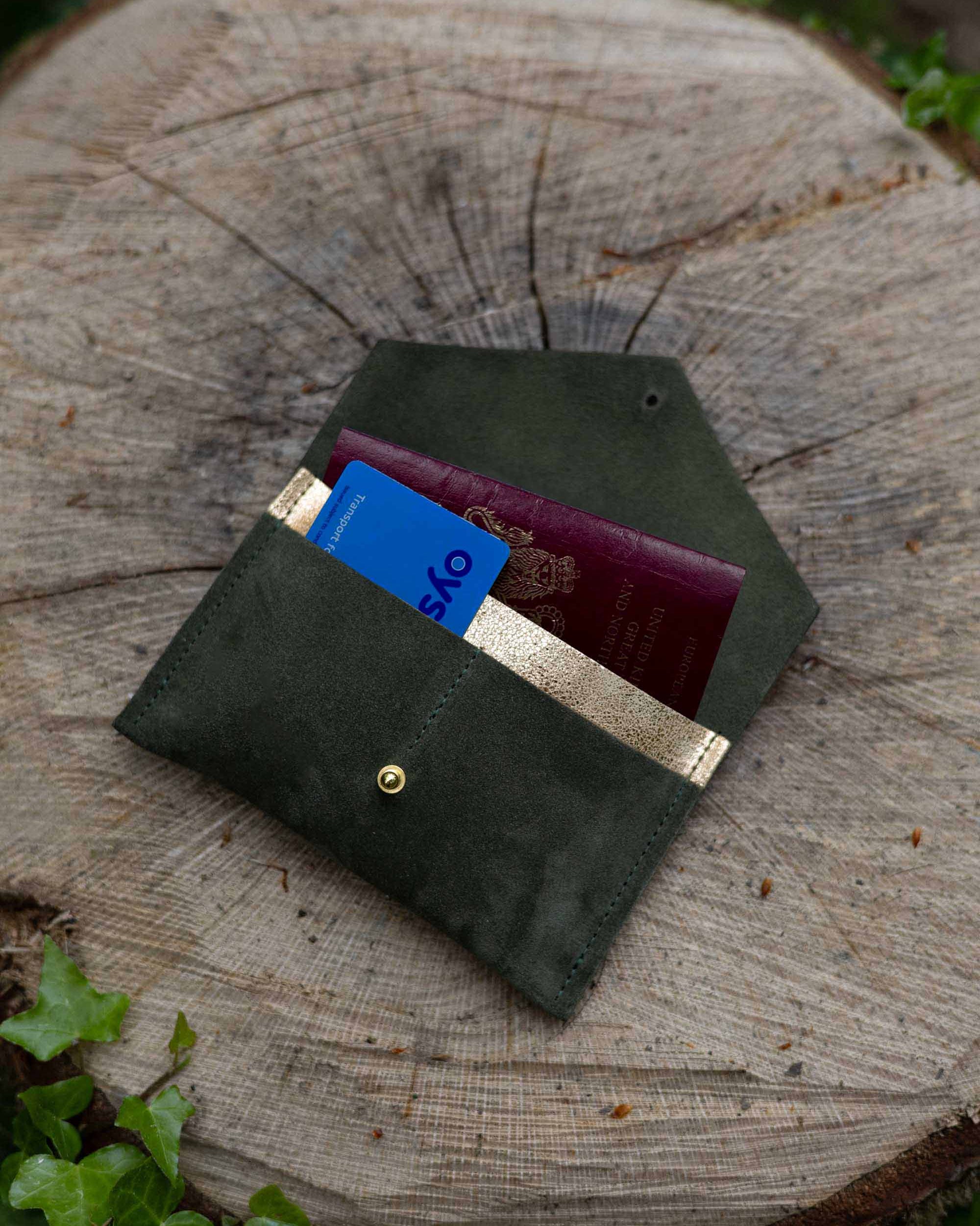 An olive green leather passport holder or card case with a gold button, shown open with a blue card and a burgundy passport inside, resting on a rustic tree stump outdoors.