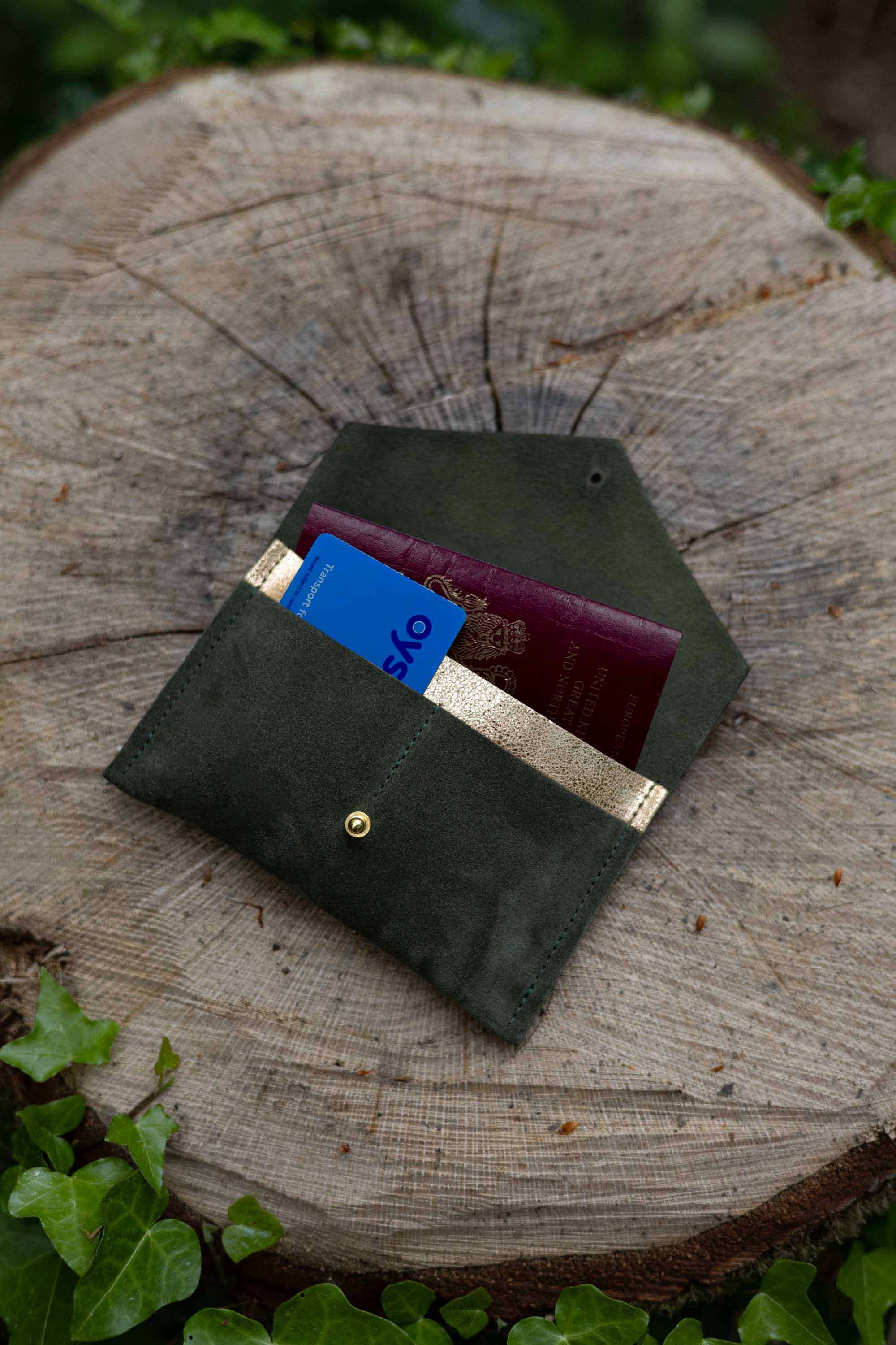 An olive green leather passport holder or card case with a gold button, shown open with a blue card and a burgundy passport inside, resting on a rustic tree stump outdoors.