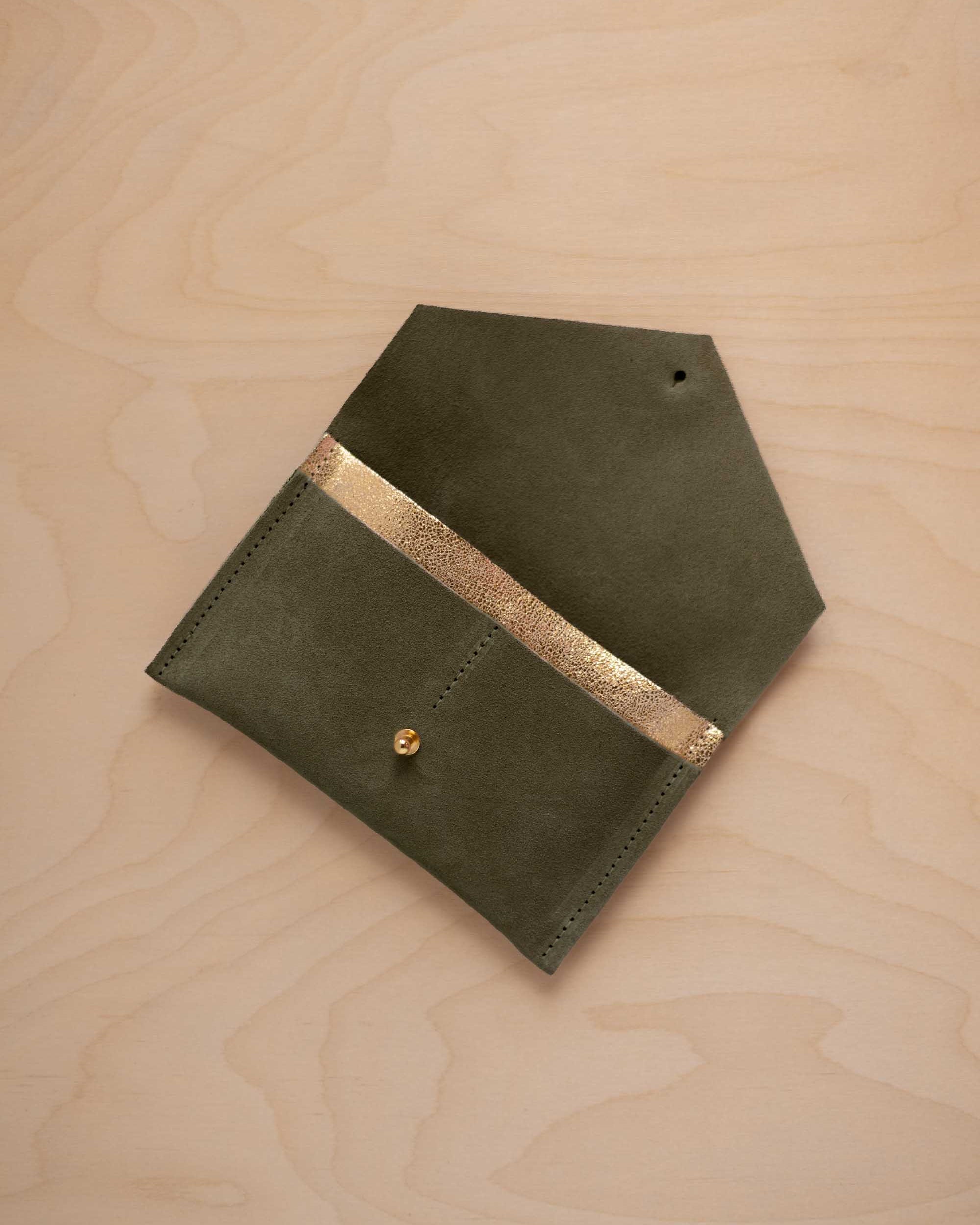 A soft olive green coloured suede pouch with a metallic gold contrast pocket and a small gold stud, displayed on a light wood grain background.