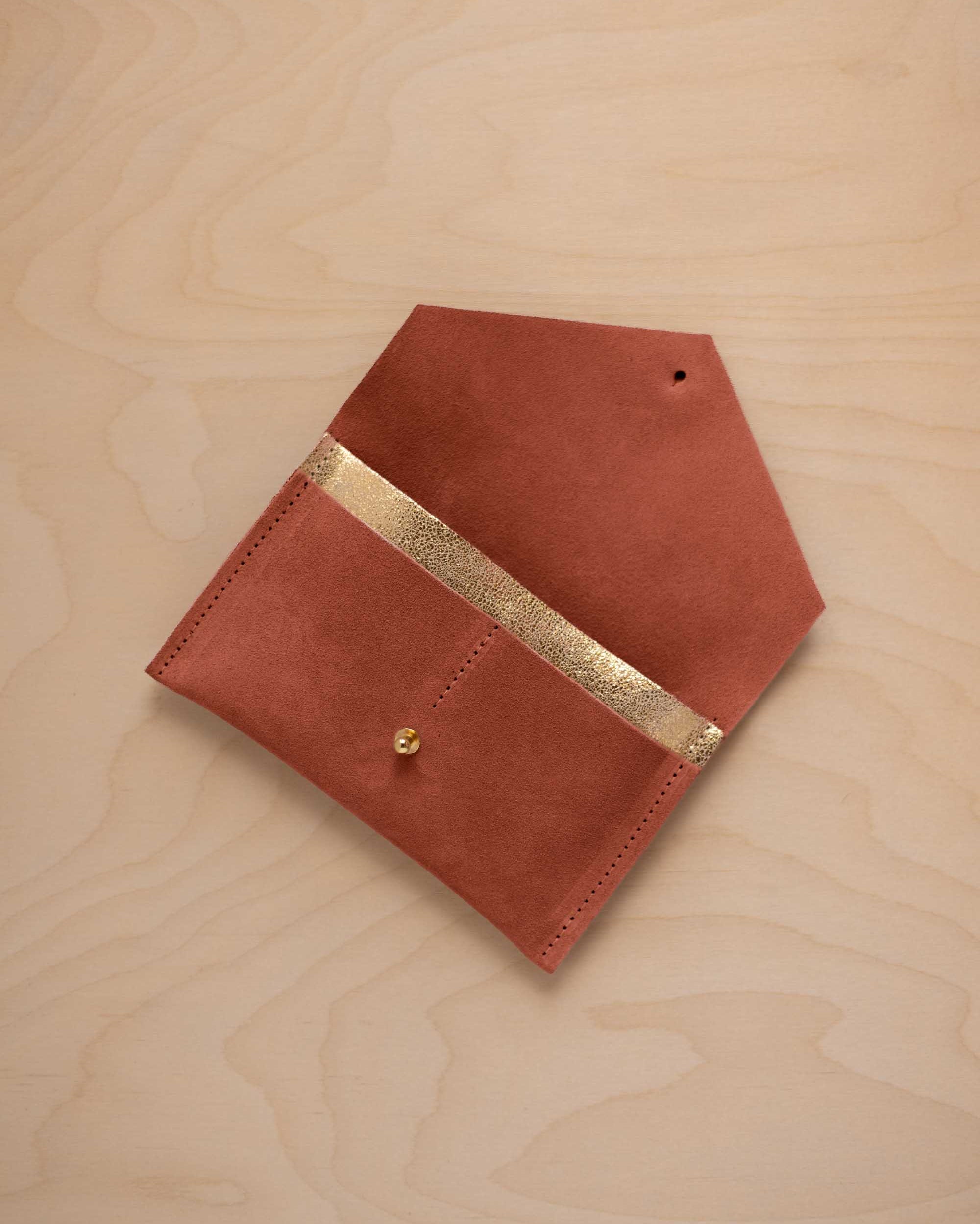 A terracotta suede pouch with a metallic gold contrast pocket and a small gold stud, displayed on a light wood grain background.