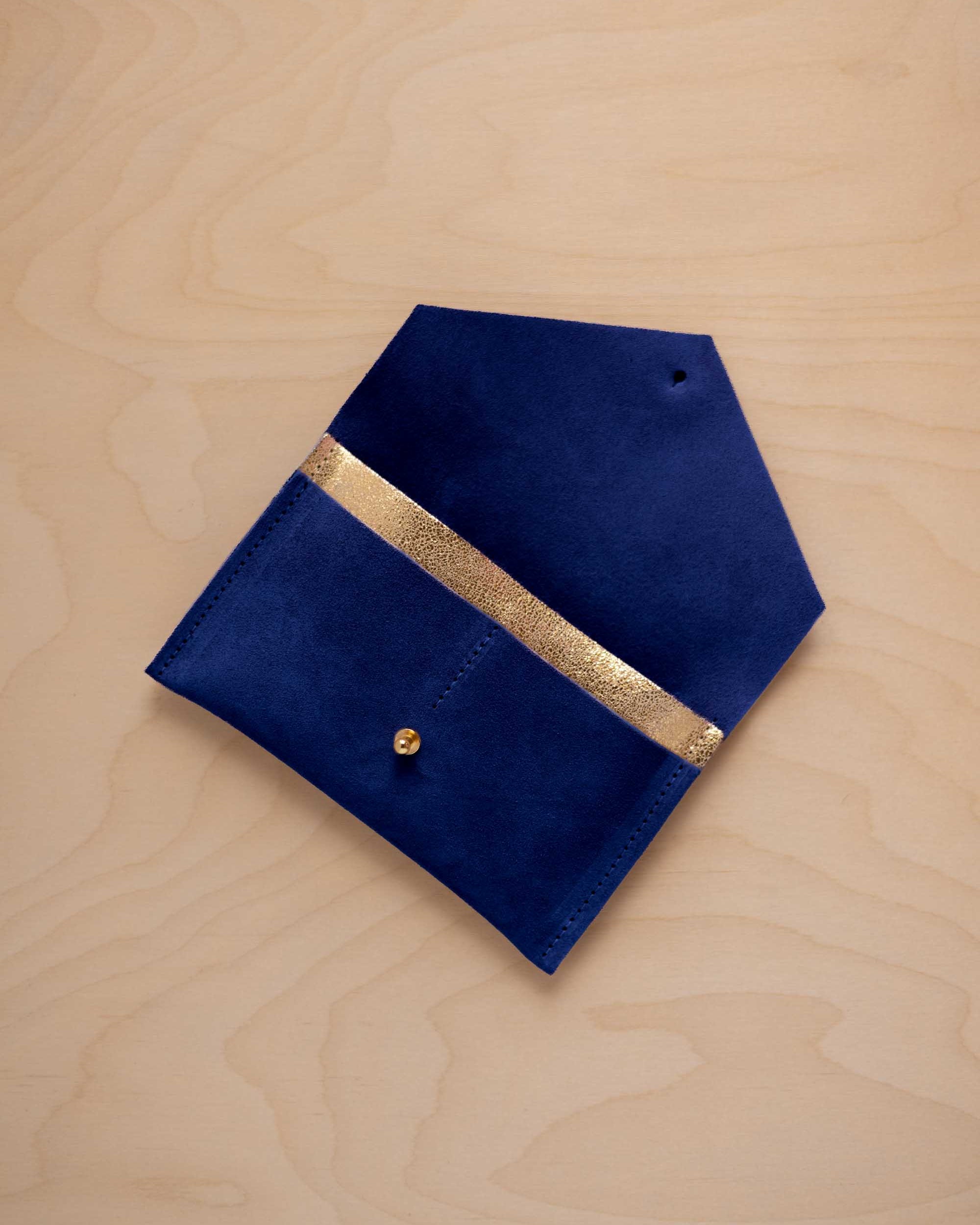 An ultramarine blue suede pouch with a metallic gold contrast pocket and a small gold stud, displayed on a light wood grain background.
