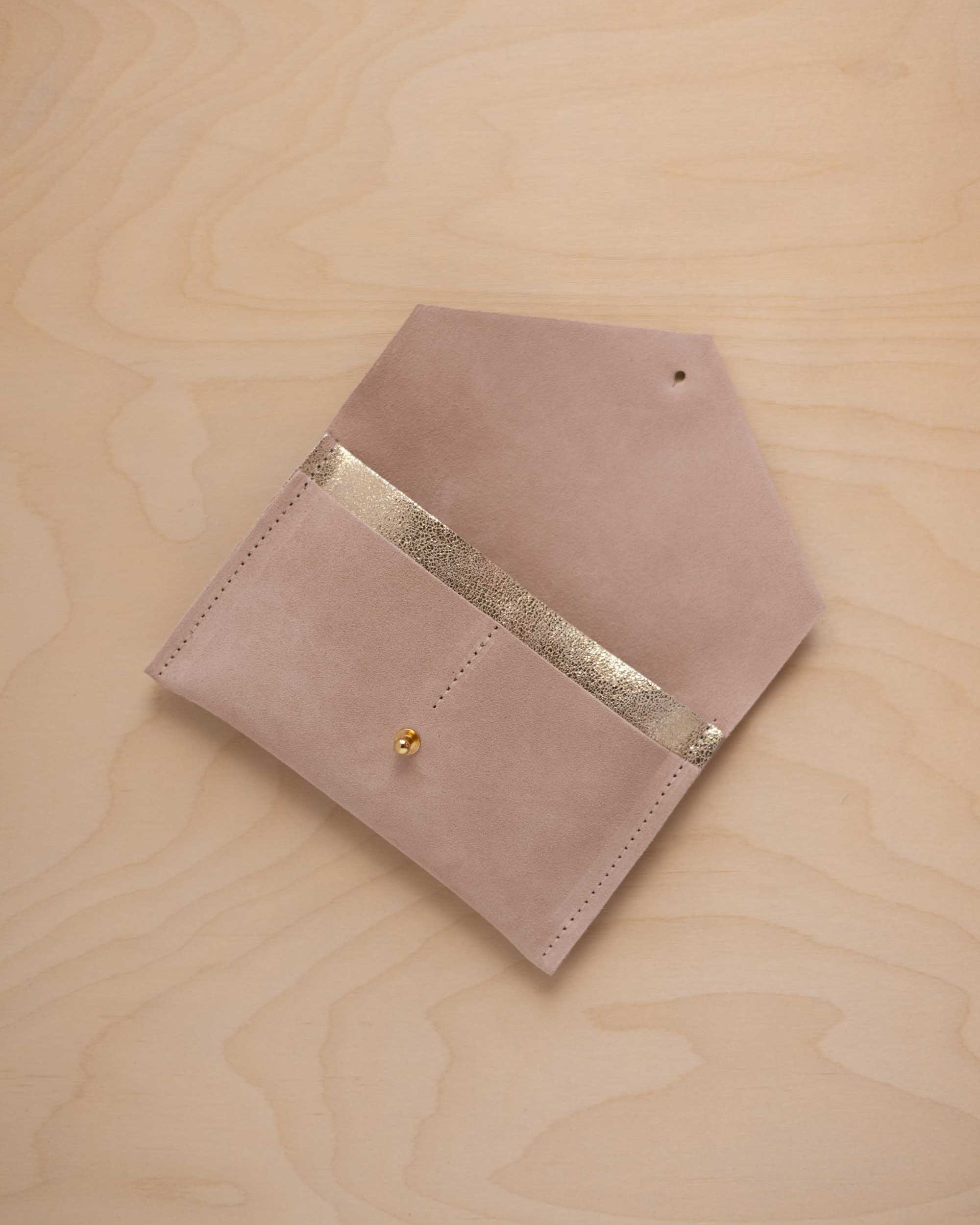 A warm beige suede pouch with a metallic gold contrast pocket and a small gold stud, displayed on a light wood grain background.