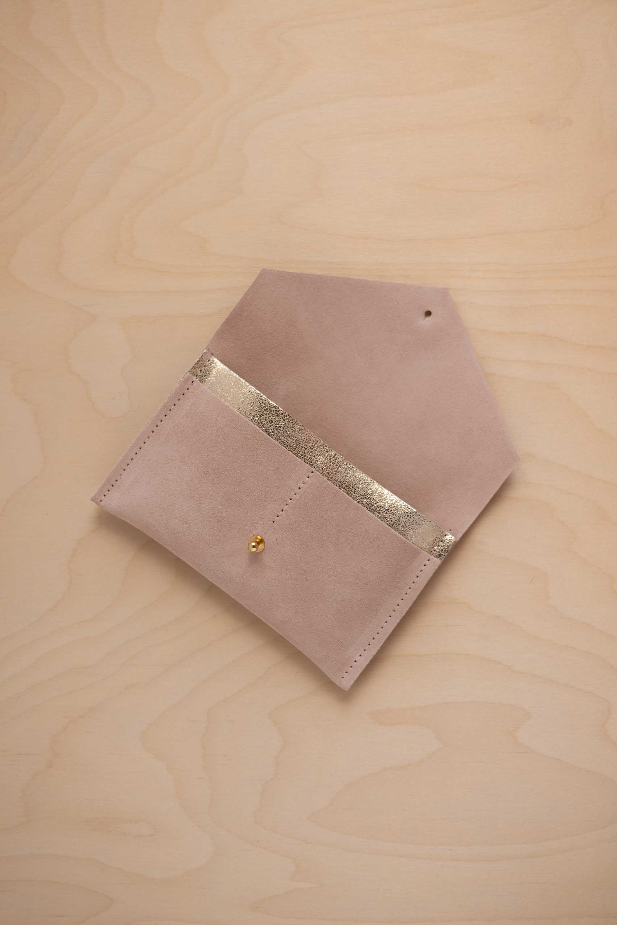 A warm beige suede pouch with a metallic gold contrast pocket and a small gold stud, displayed on a light wood grain background.