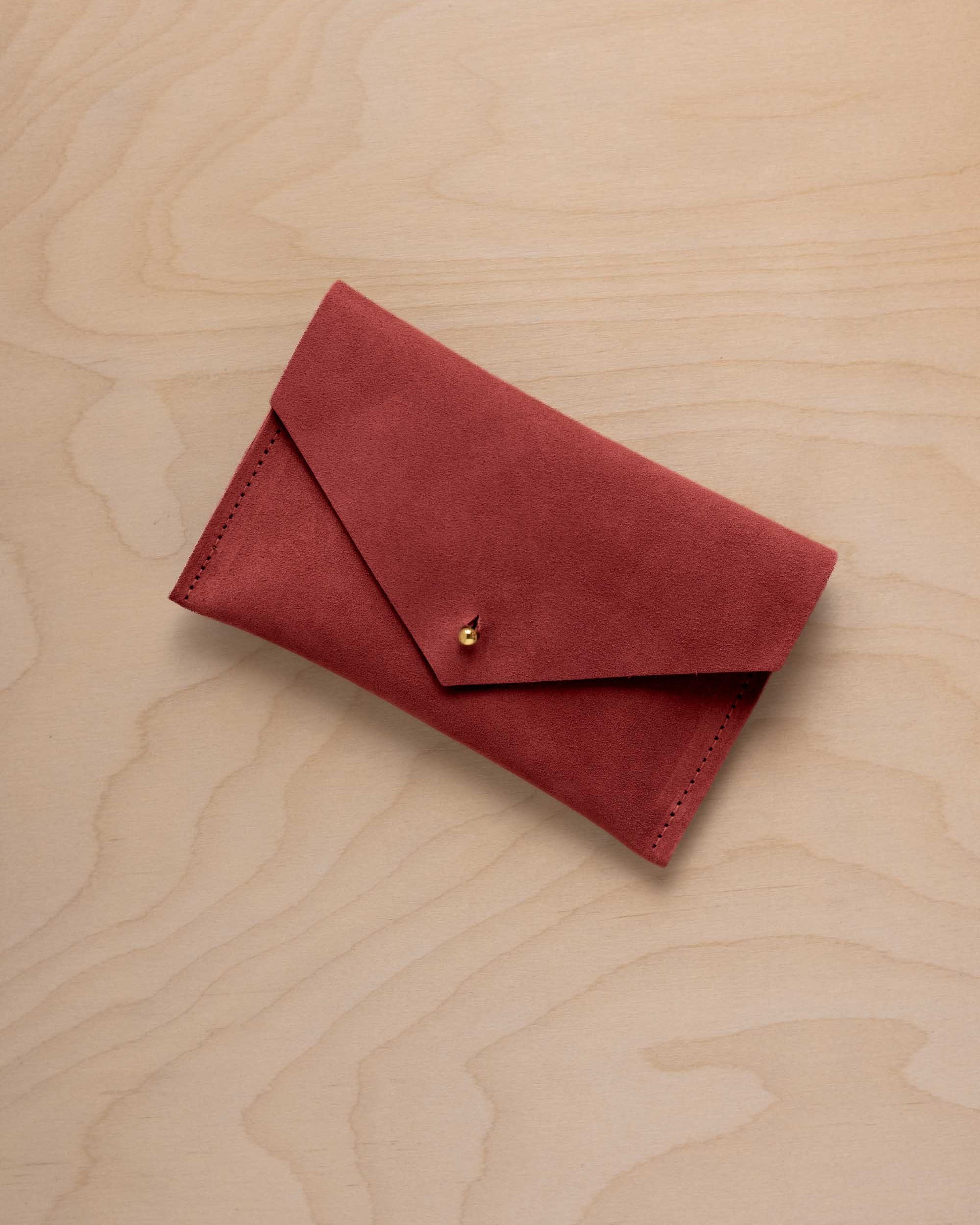 A small red suede envelope-style pouch with a gold snap button, placed on a light wood grain surface.