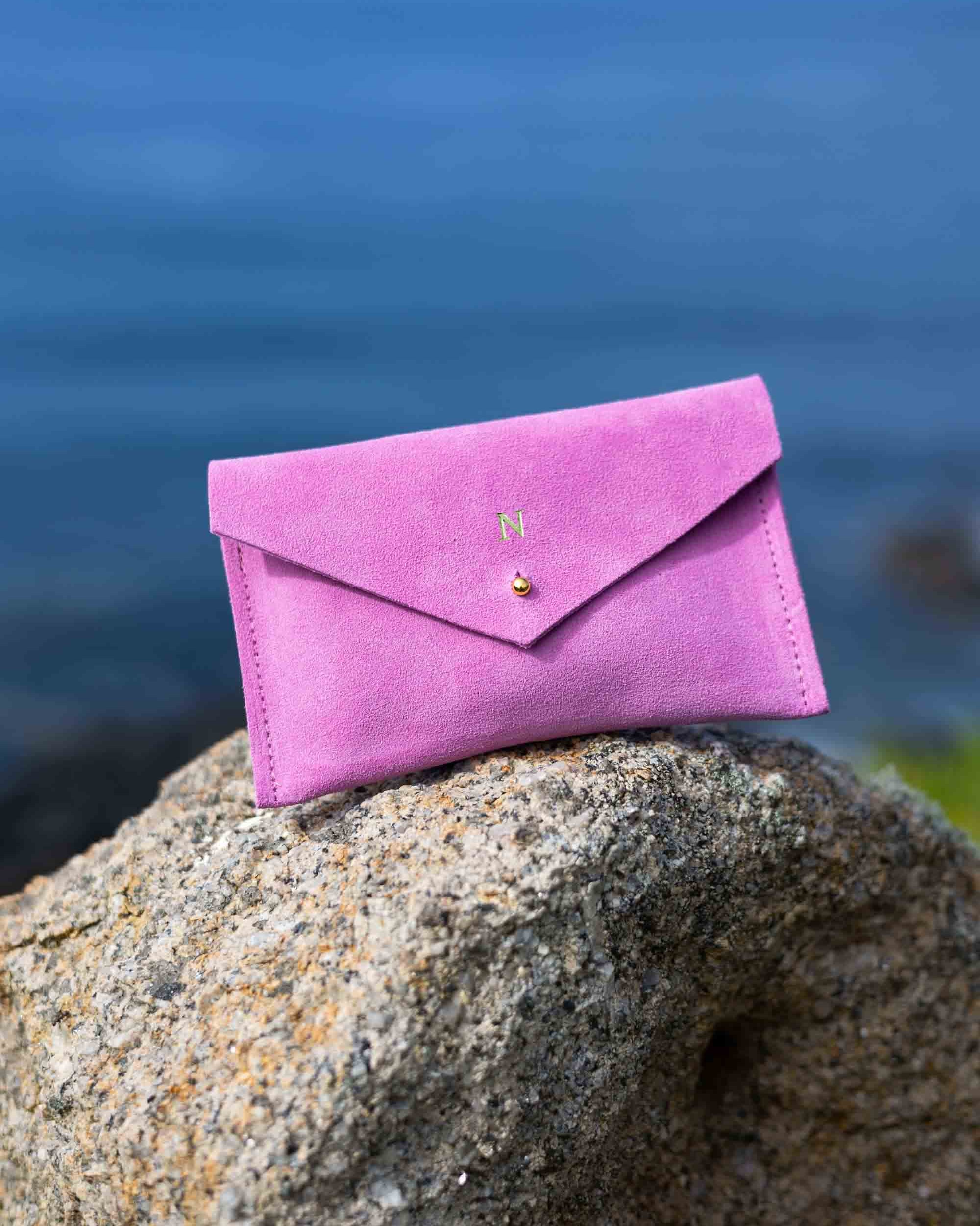 A small peony pink suede envelope-style pouch with a gold foiled initial and gold button, resting on a large textured rock with a blurred outdoor background.