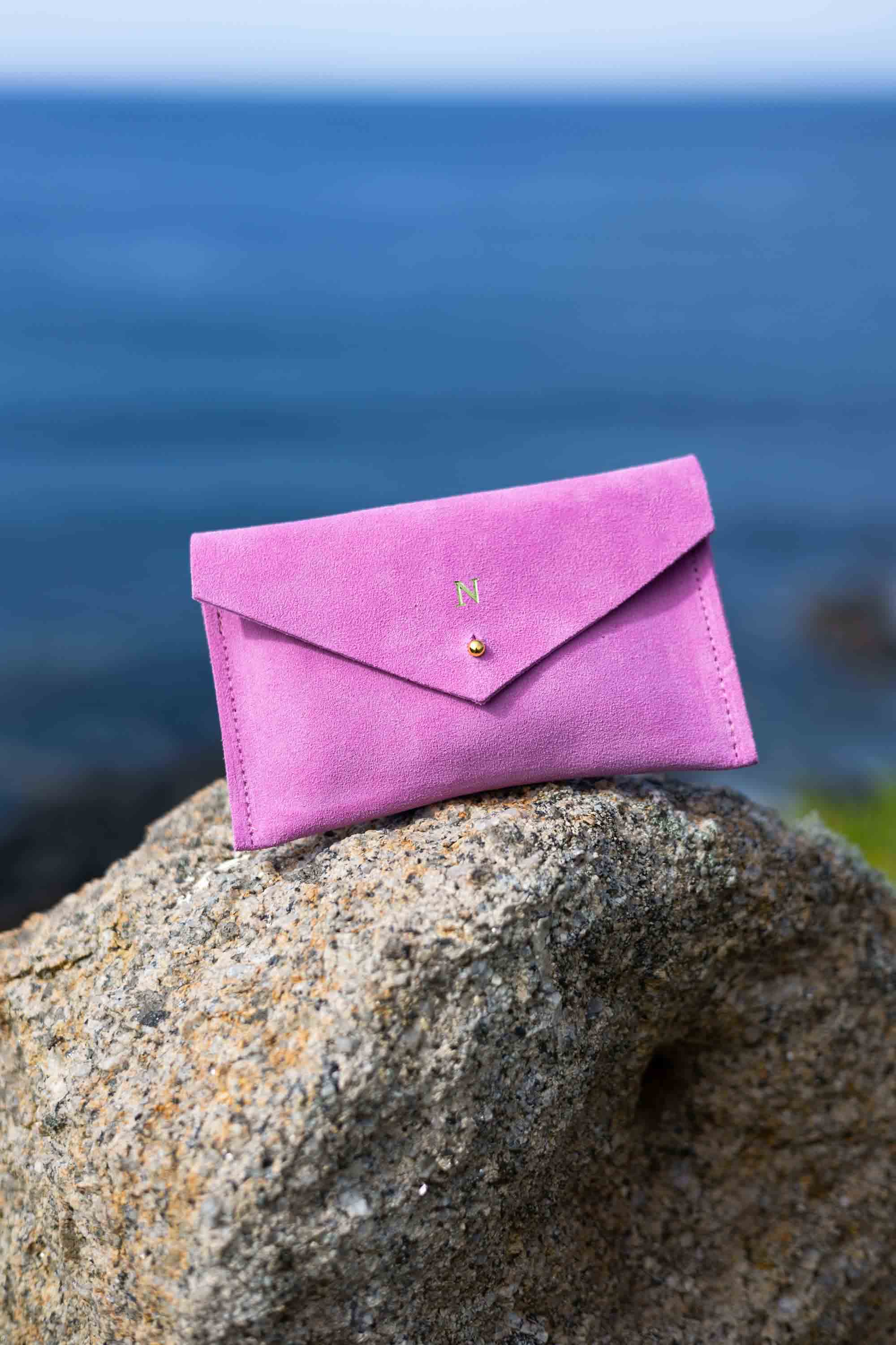 A small peony pink suede envelope-style pouch with a gold foiled initial and gold button, resting on a large textured rock with a blurred outdoor background.