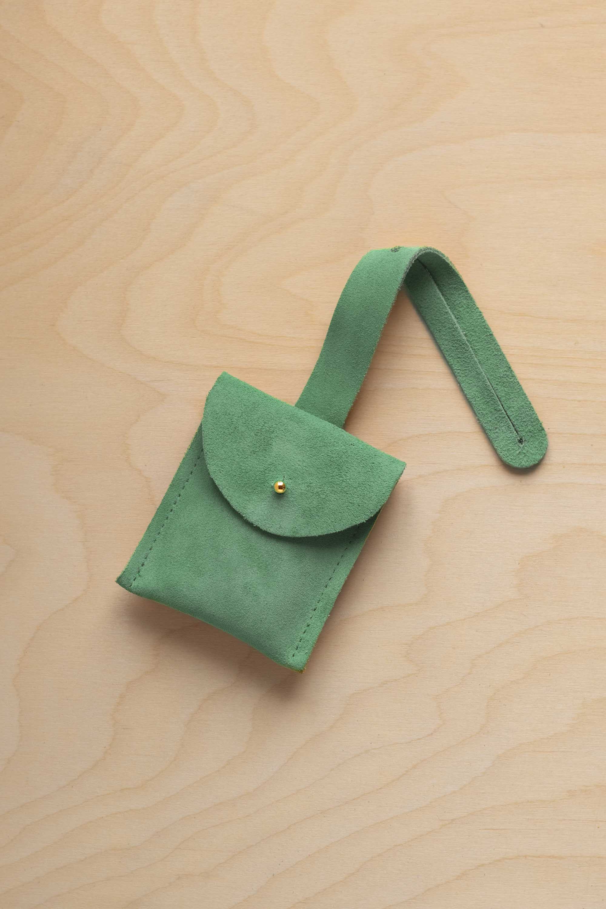 A small seafoam turquoise green suede pouch with a flap closure and a matching strap, resting on a light wood grain surface.