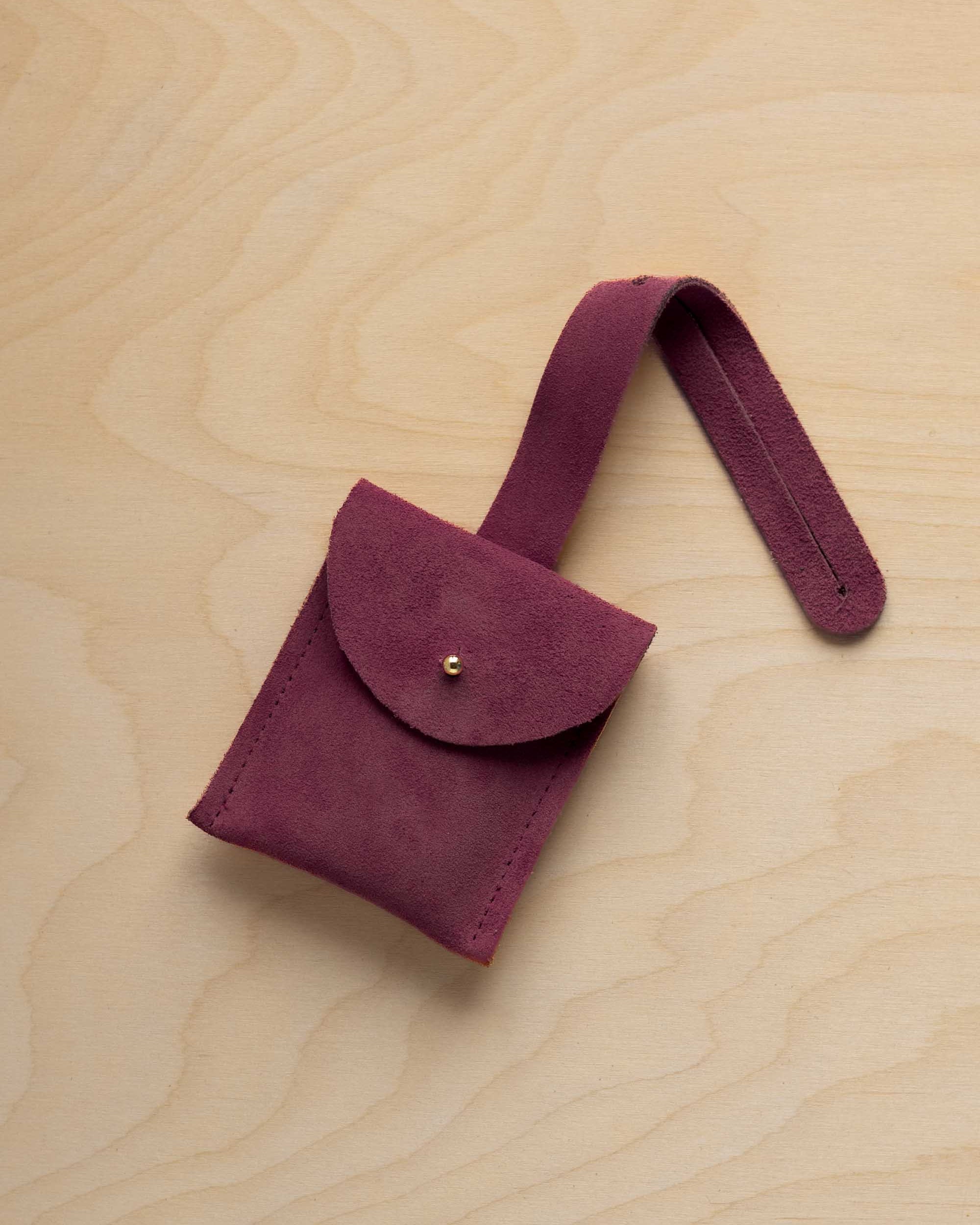 A small burgundy suede pouch with a flap closure and a matching strap, resting on a light wood grain surface.
