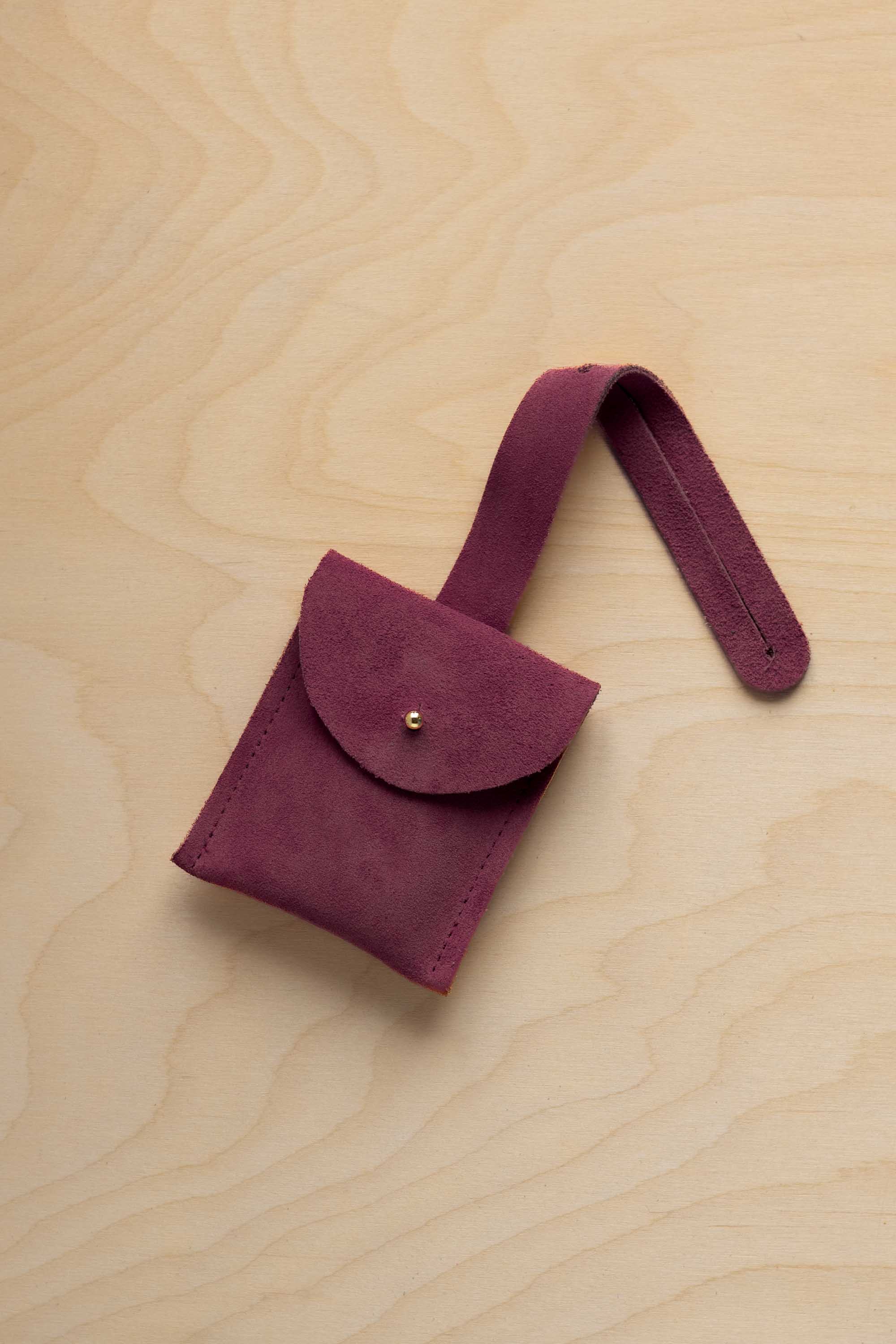 A small burgundy suede pouch with a flap closure and a matching strap, resting on a light wood grain surface.