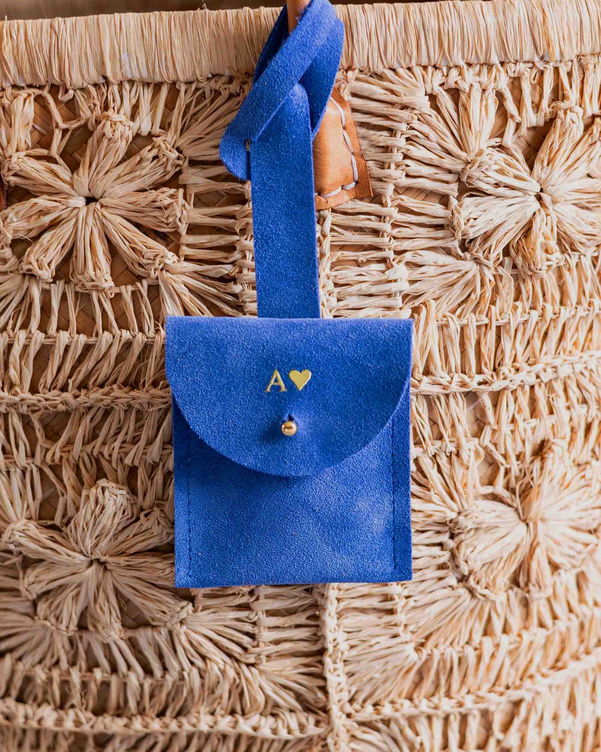 A personalised blue suede luggage tag pouch with the intial "A" on it, a strap and gold button, hanging from a leather handle on a woven straw bag.