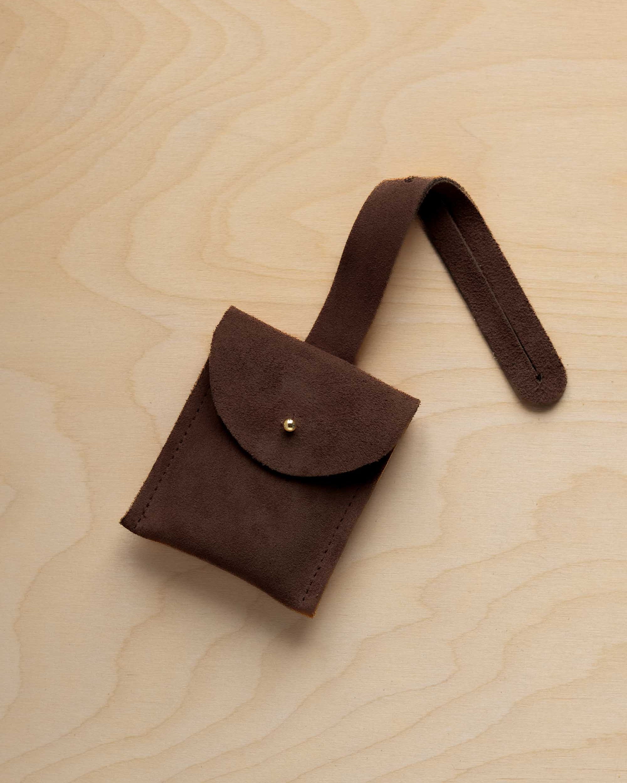 A small espresso brown suede pouch with a flap closure and a matching strap, resting on a light wood grain surface.