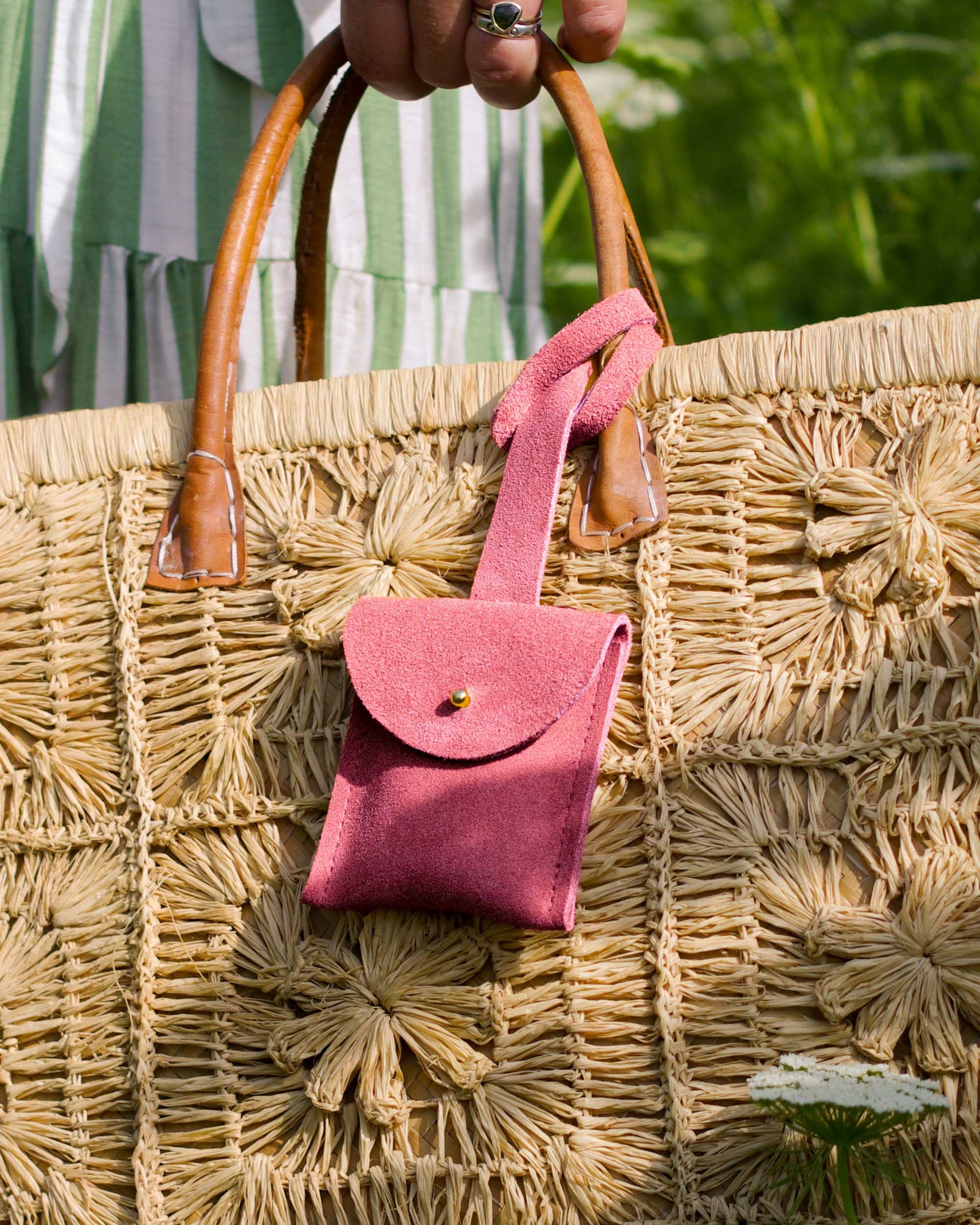 A close-up of a large woven straw tote bag with brown leather handles, featuring a small pink suede luggage tag pouch hanging from one handle. A person's hand with a ring is holding the bag, and a green and white striped dress is visible in the background.
