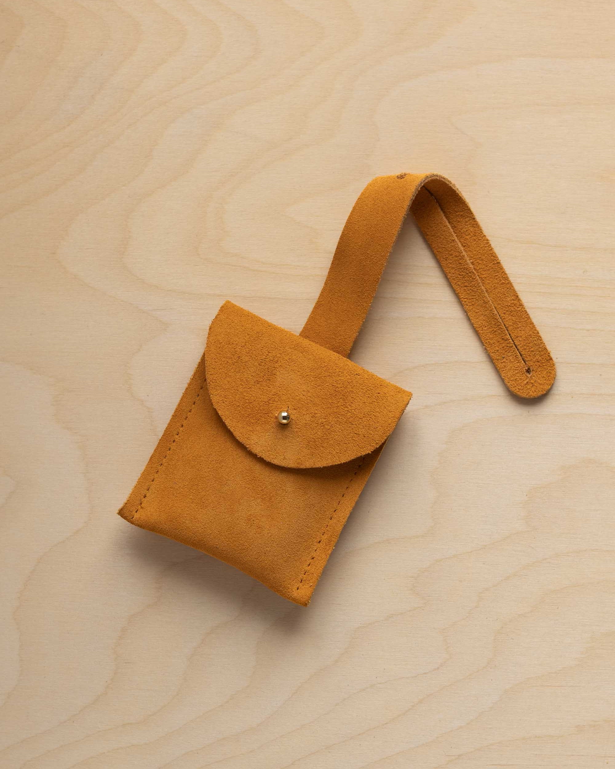 A small golden ochre mustard yellow coloured suede pouch with a flap closure and a matching strap, resting on a light wood grain surface.