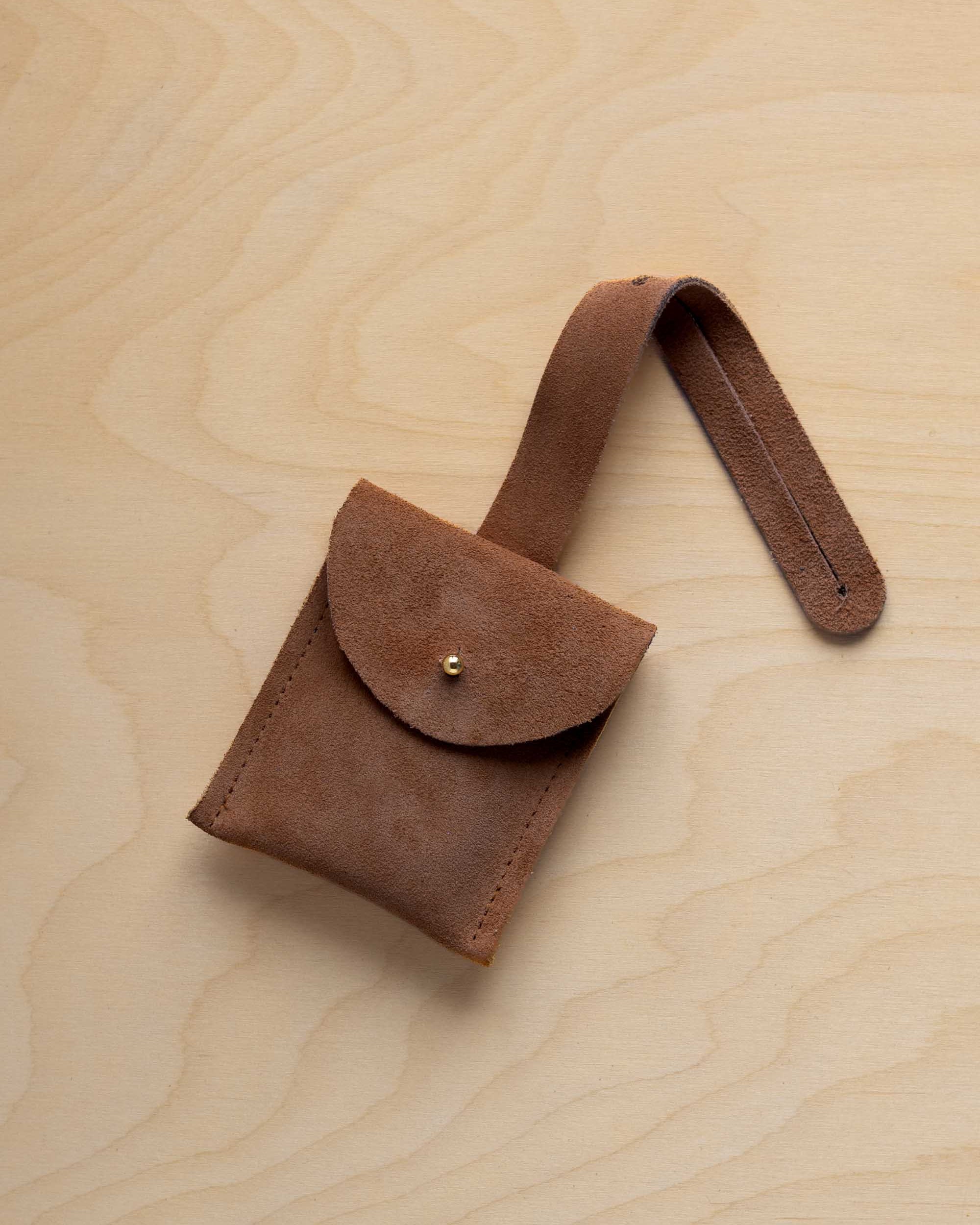A small mocha brown suede pouch with a flap closure and a matching strap, resting on a light wood grain surface.