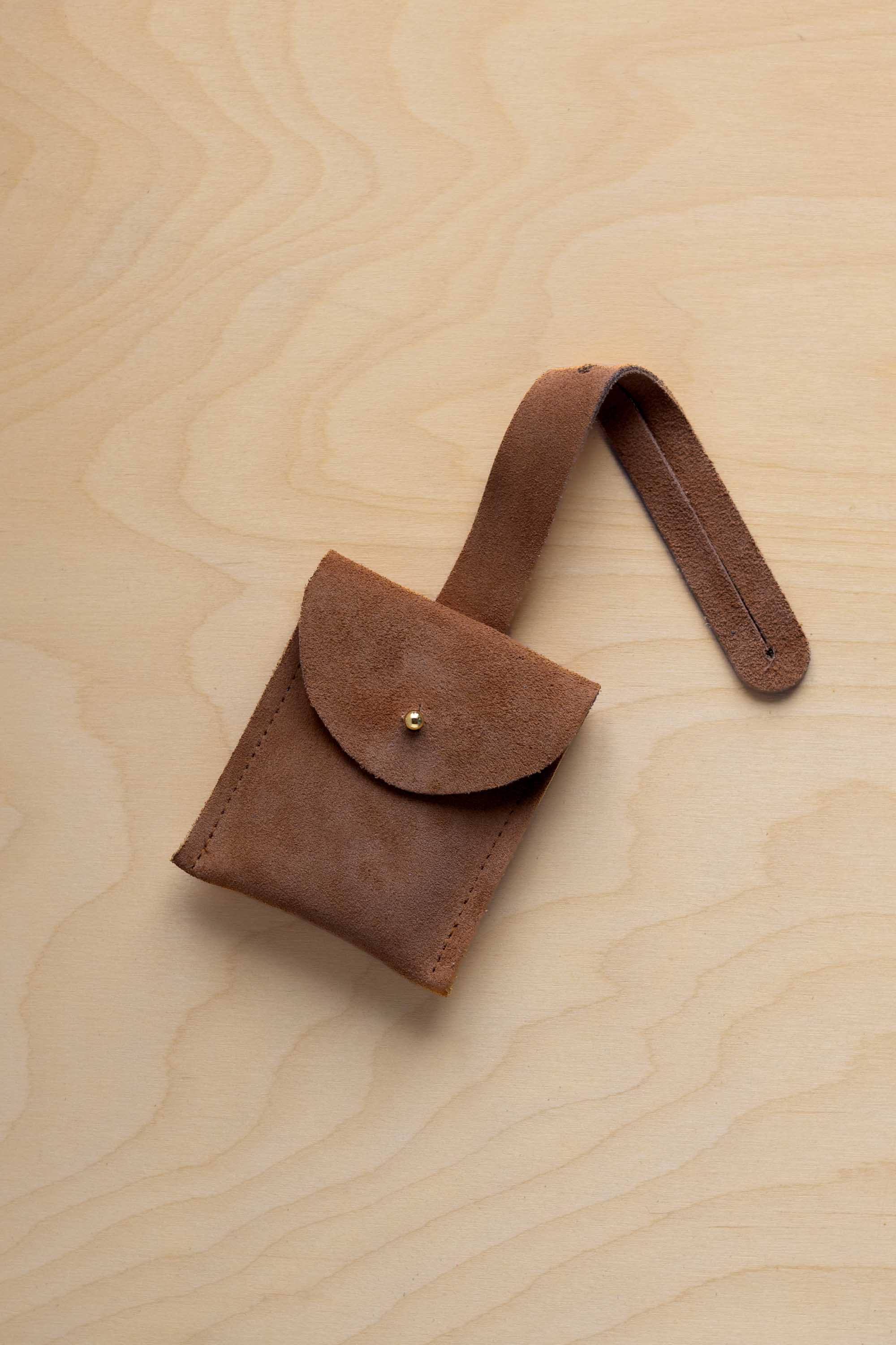 A small mocha brown suede pouch with a flap closure and a matching strap, resting on a light wood grain surface.