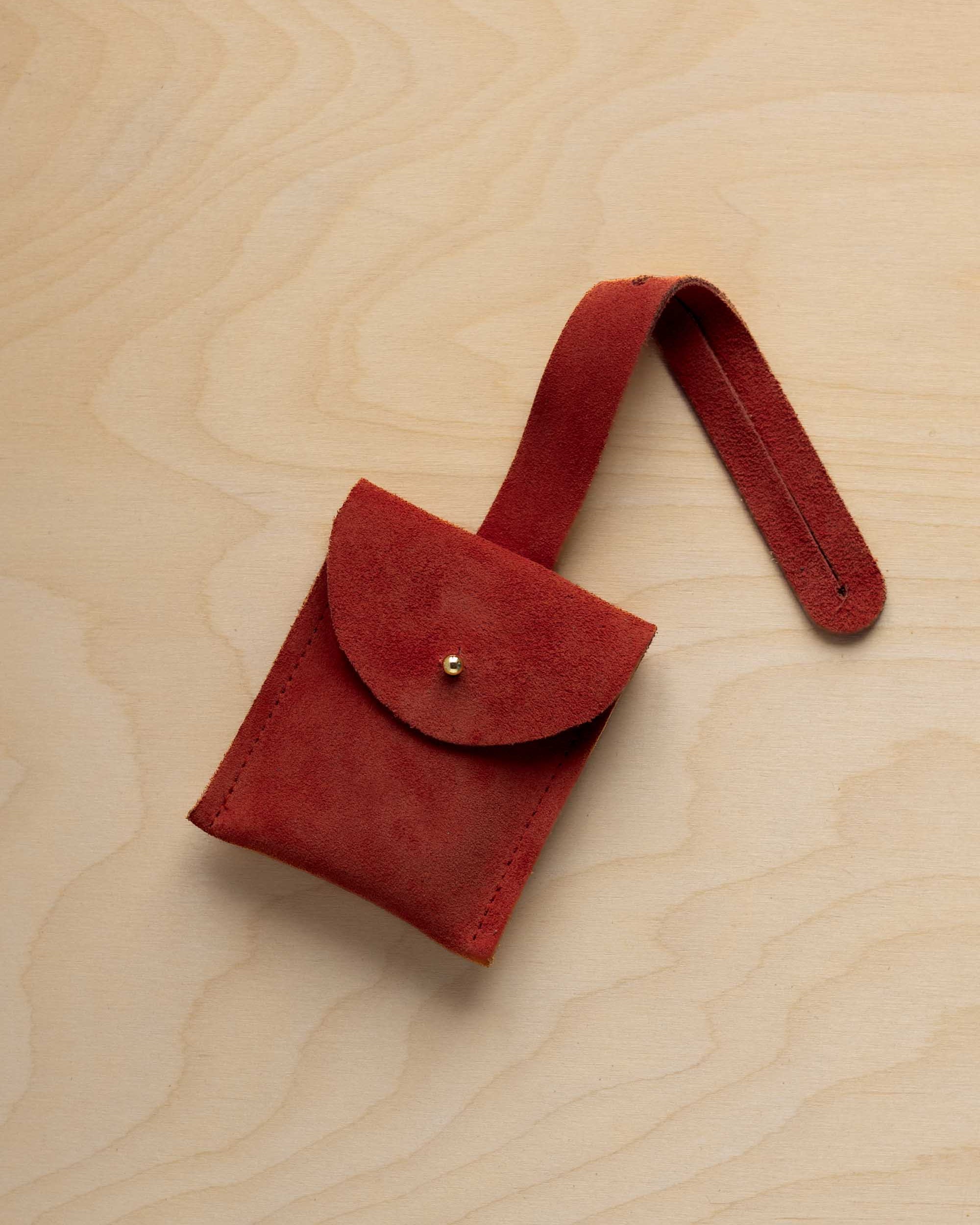 A small rust red coloured suede pouch with a flap closure and a matching strap, resting on a light wood grain surface.
