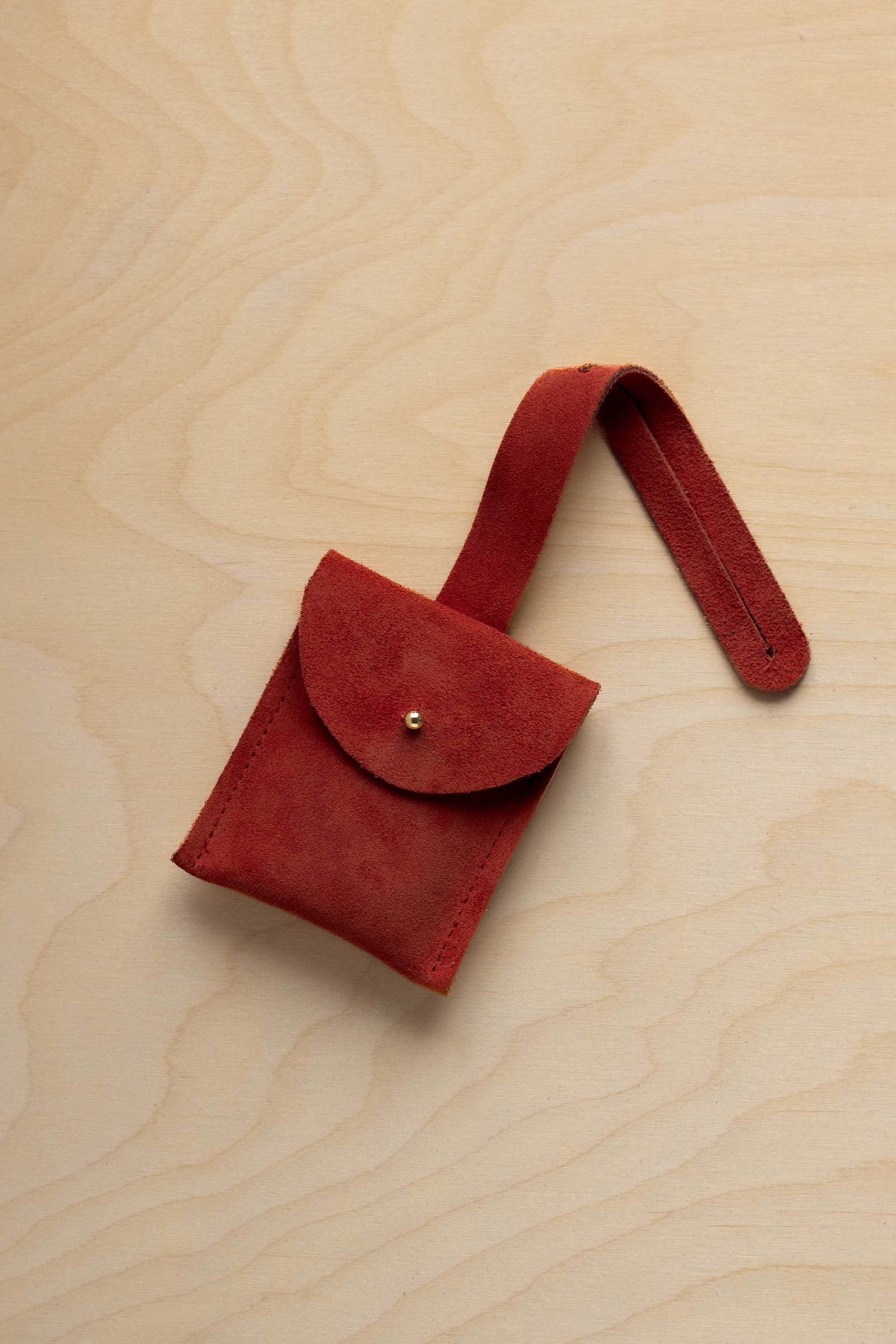 A small rust red coloured suede pouch with a flap closure and a matching strap, resting on a light wood grain surface.