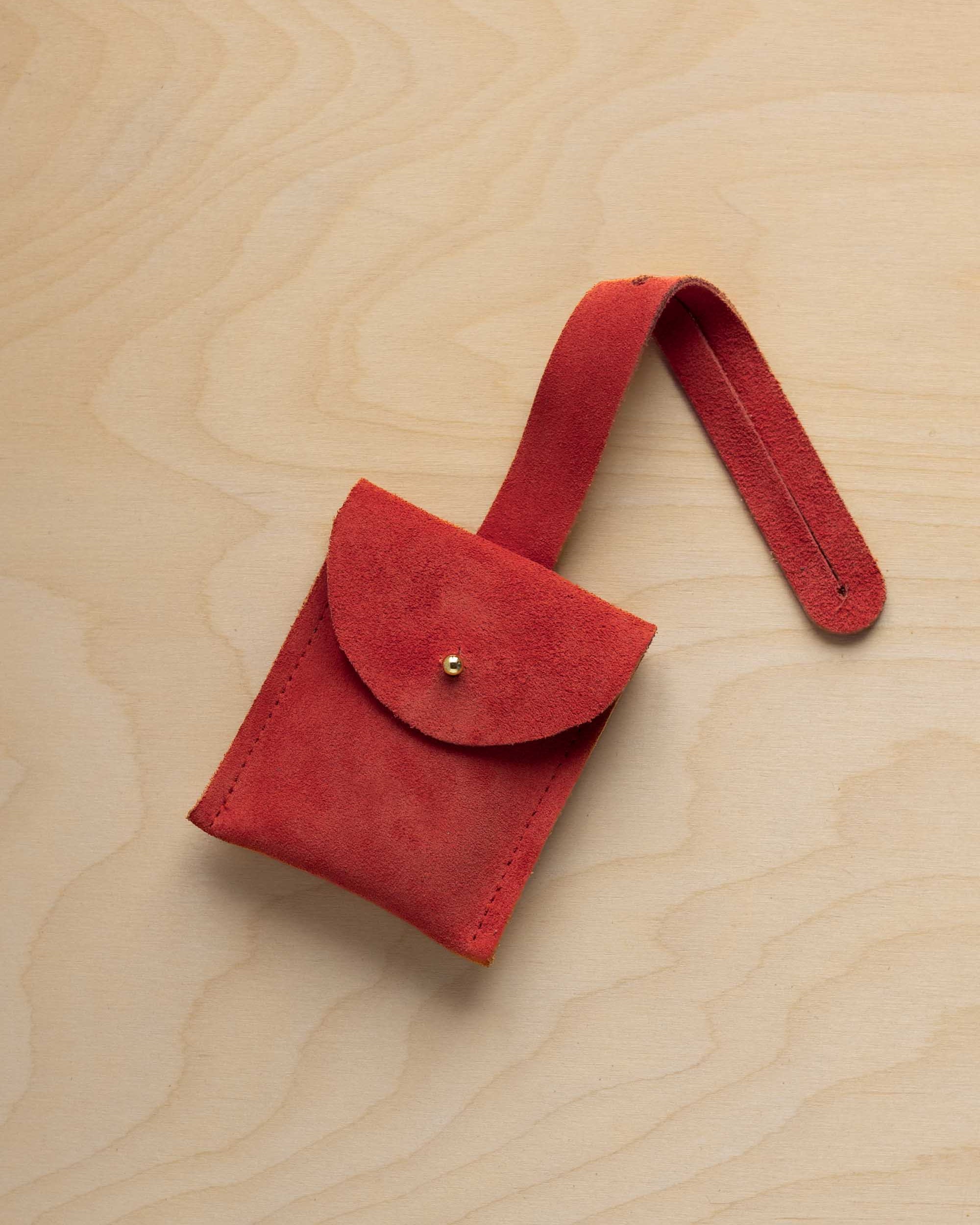 A small scarlett red coloured suede pouch with a flap closure and a matching strap, resting on a light wood grain surface.