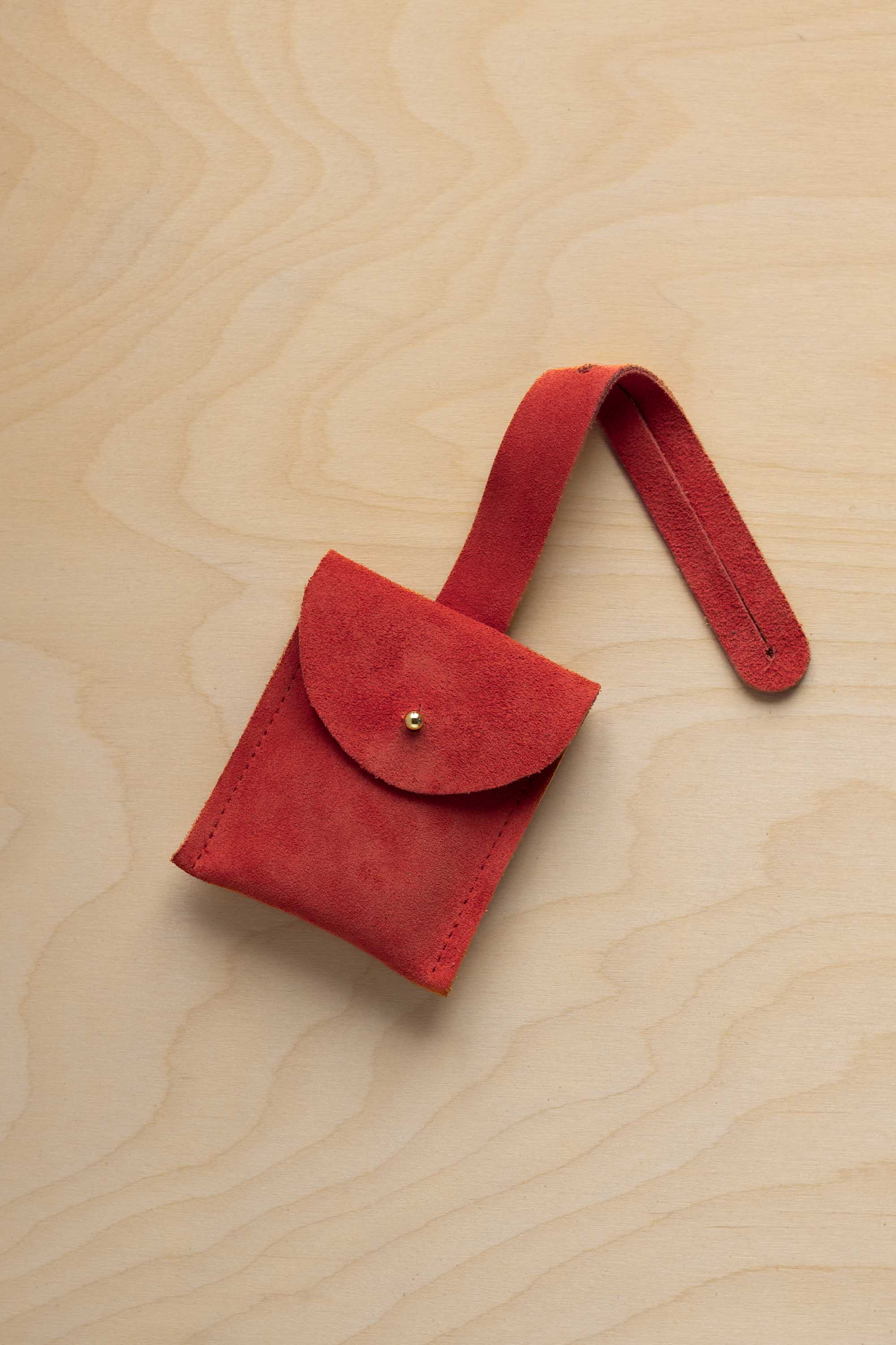 A small scarlett red coloured suede pouch with a flap closure and a matching strap, resting on a light wood grain surface.
