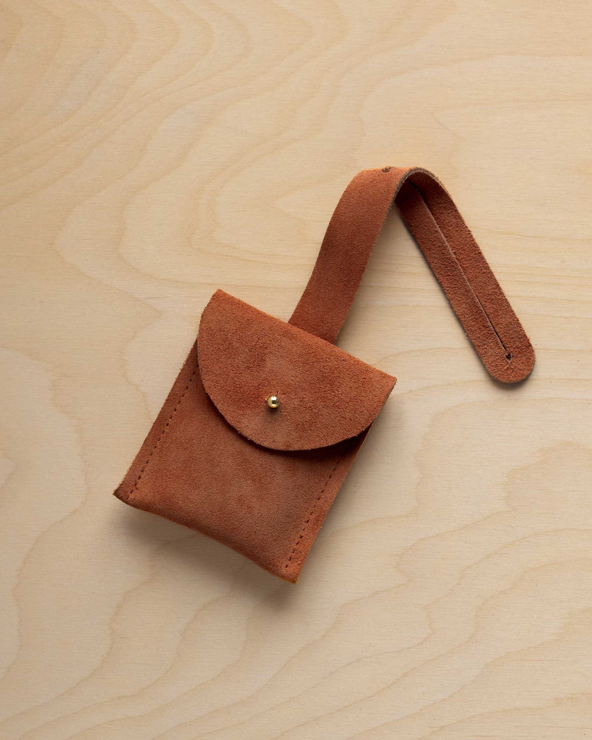 A small terracotta coloured suede pouch with a flap closure and a matching strap, resting on a light wood grain surface.