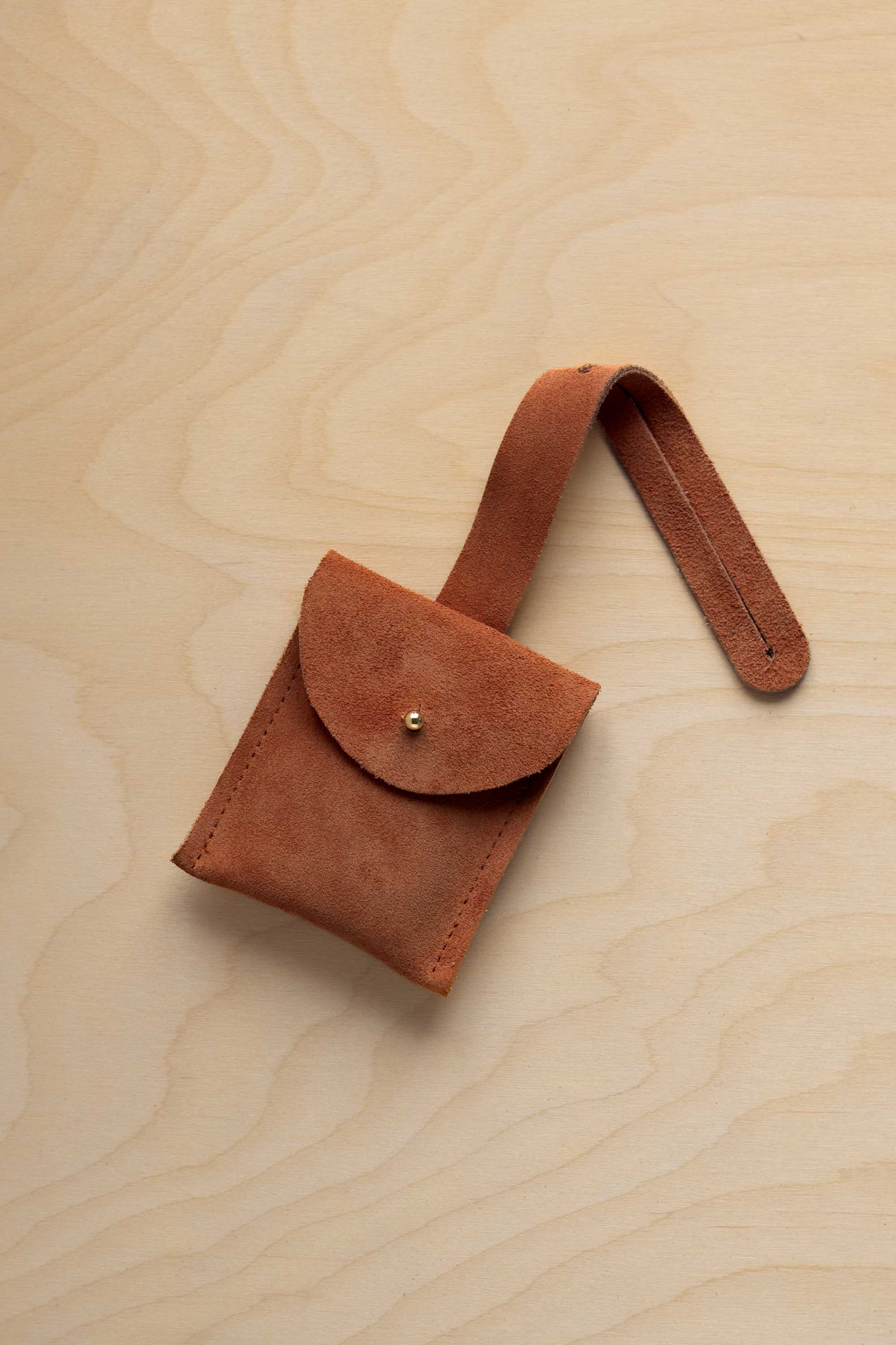 A small terracotta coloured suede pouch with a flap closure and a matching strap, resting on a light wood grain surface.