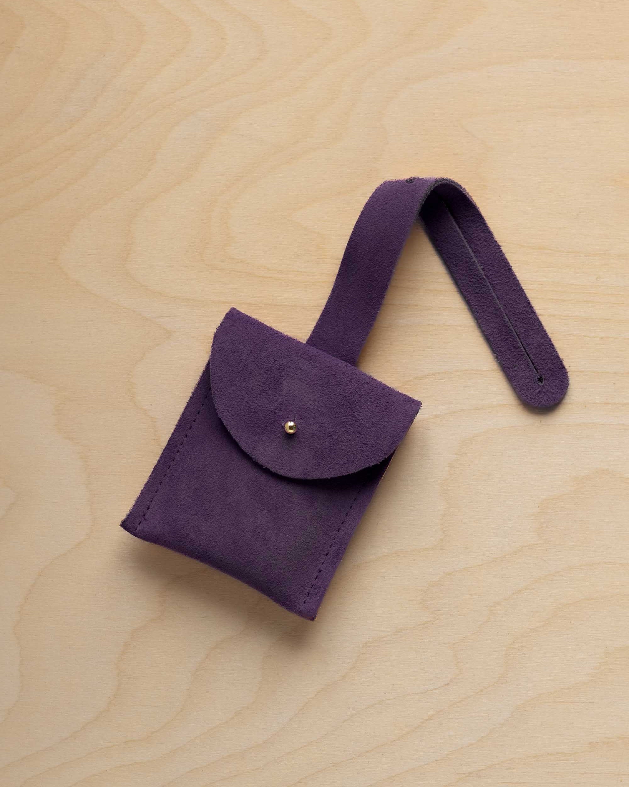 A small blackcurrant dark purple coloured suede pouch with a flap closure and a matching strap, resting on a light wood grain surface.