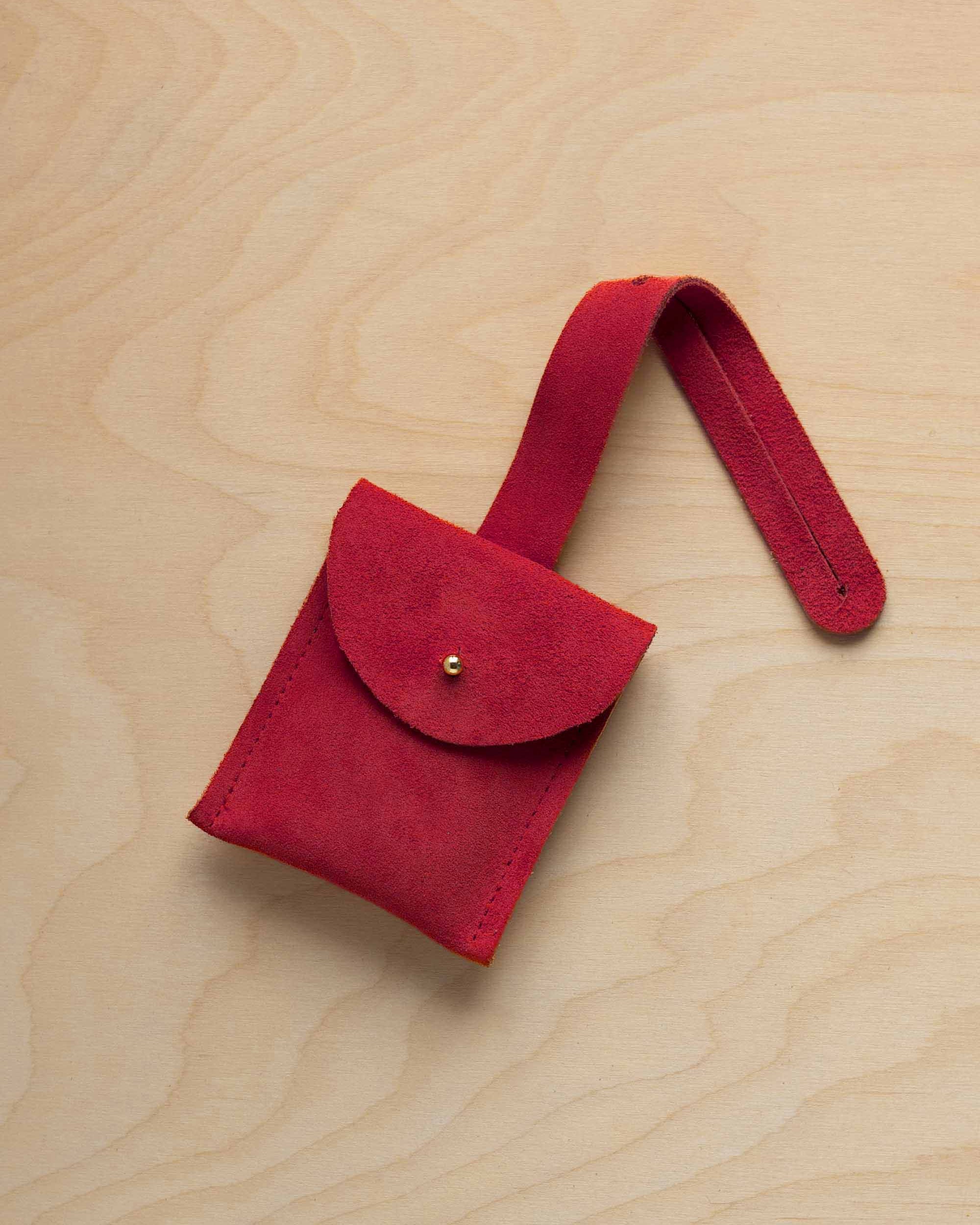 A small crimson red coloured suede pouch with a flap closure and a matching strap, resting on a light wood grain surface.